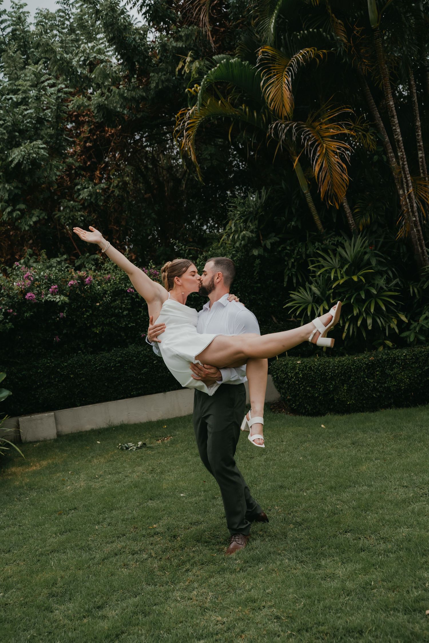 Groom lifts bride in arms as she extends leg gracefully in a fun outdoor wedding portrait in tropical garden.
