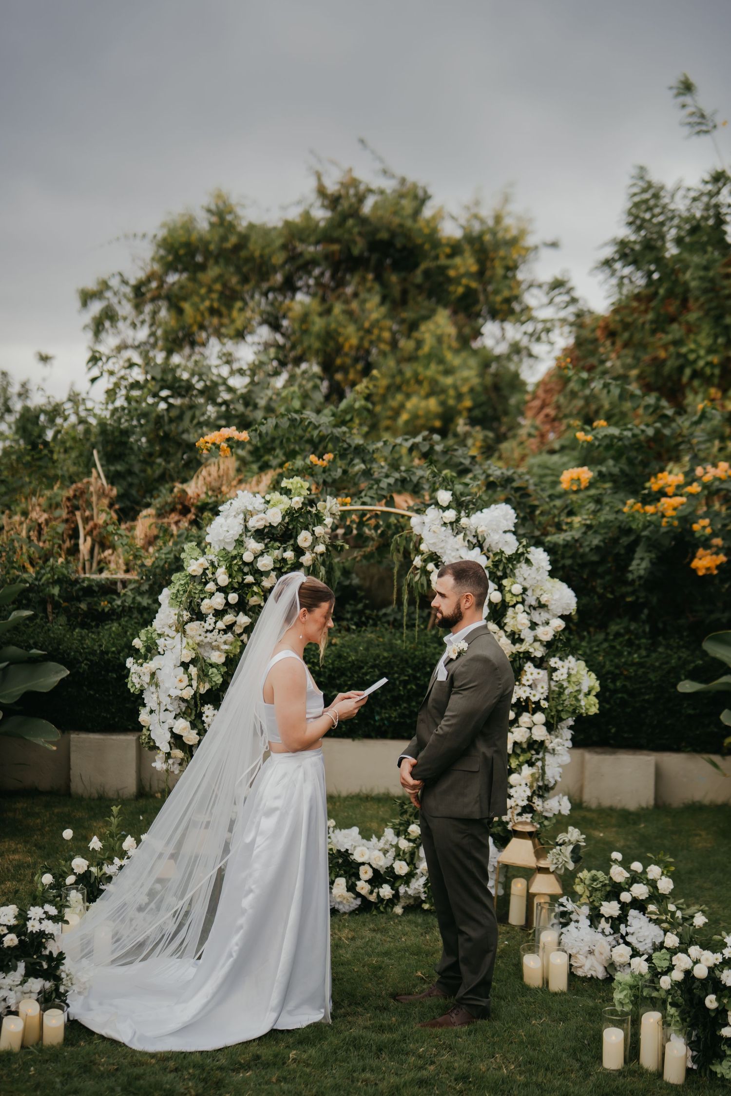 Bride reading vows to groom at white floral wedding arch, surrounded by candles and lush garden greenery.