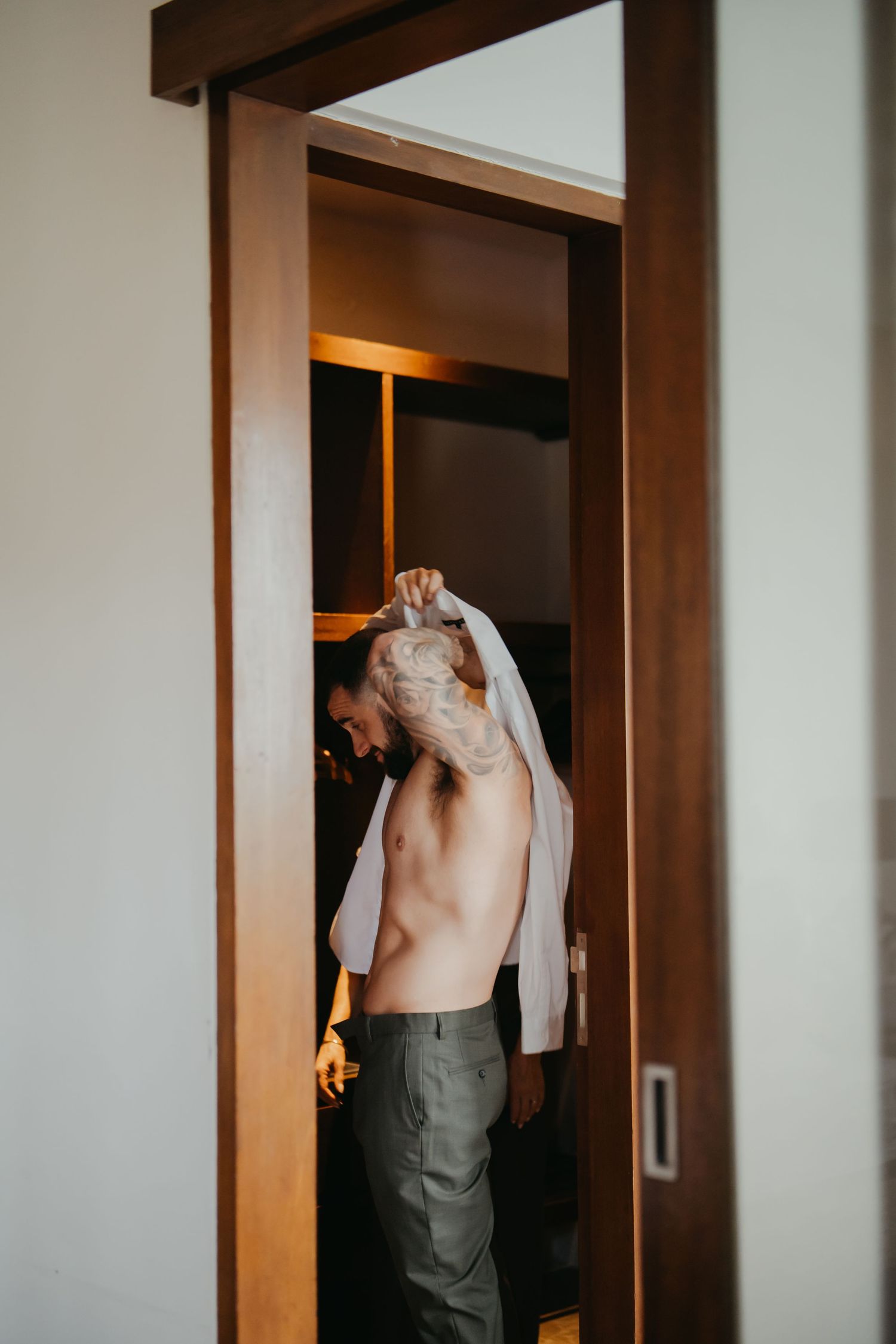 Groom putting on his white dress shirt before wedding ceremony in a warmly lit hotel room.