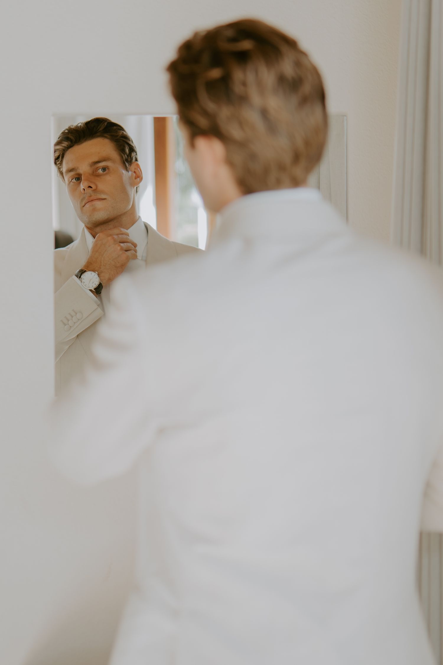 Handsome groom in white suit adjusting his collar while looking at his reflection in a mirror.
