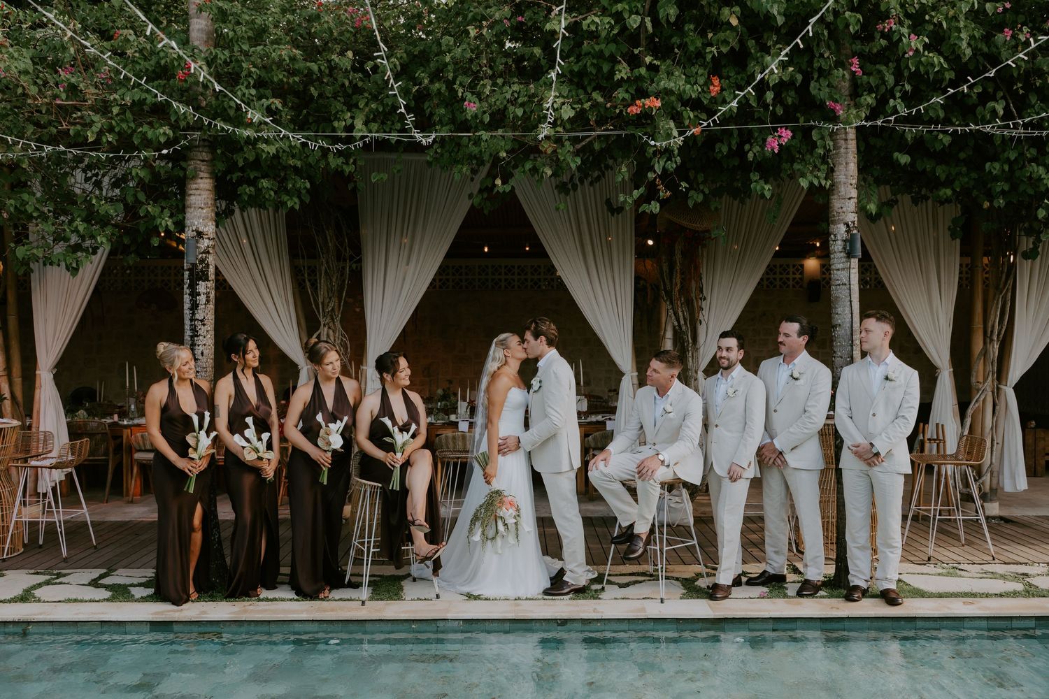 Wedding party poses poolside with bridesmaids in black and groomsmen in white suits under trees.