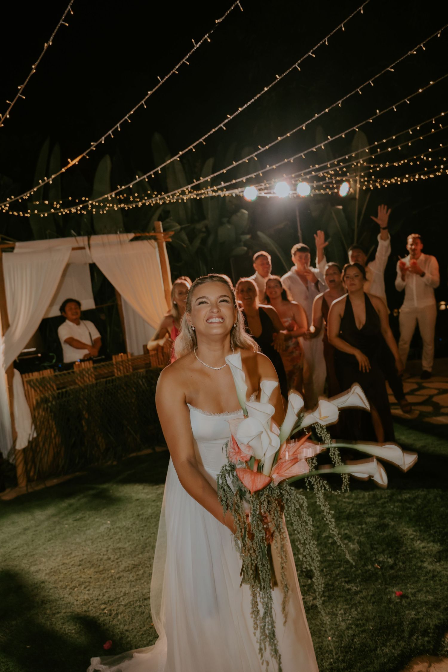 Bride smiles while preparing to toss her bouquet at an outdoor night wedding reception with string lights.