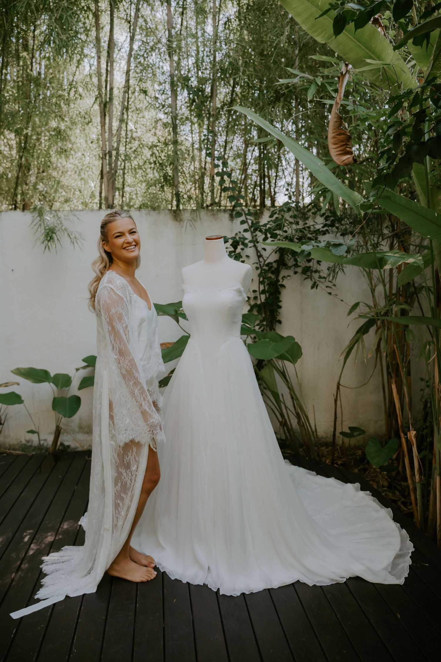 Smiling bride in lace robe stands beside wedding dress on mannequin on outdoor deck surrounded by lush greenery.