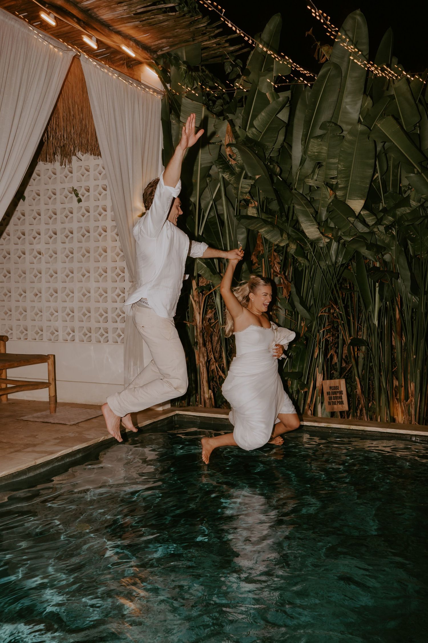 Bride and groom jump into pool together at night during wedding celebration.