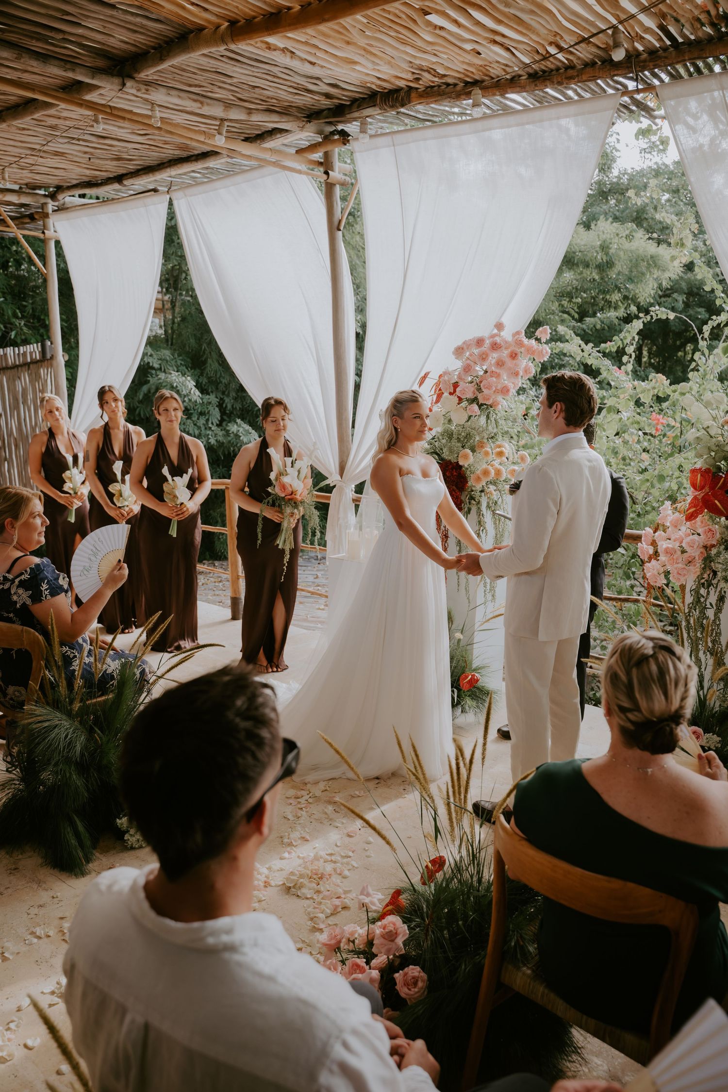 Bride and groom exchanging vows under white draped canopy with bridesmaids in dark dresses at outdoor ceremony.