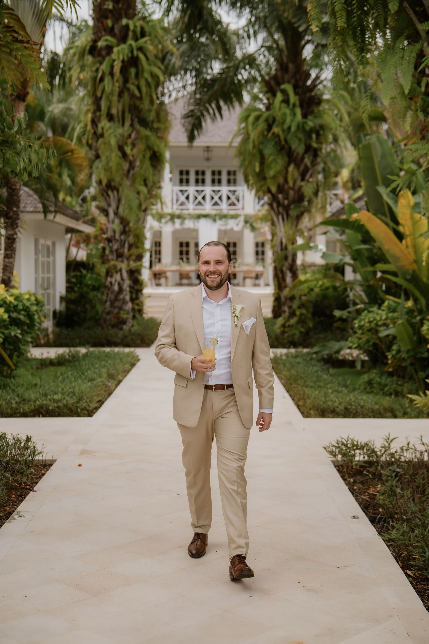 Smiling groom in a tan suit walking along a palm-lined pathway holding a cocktail at a tropical resort.