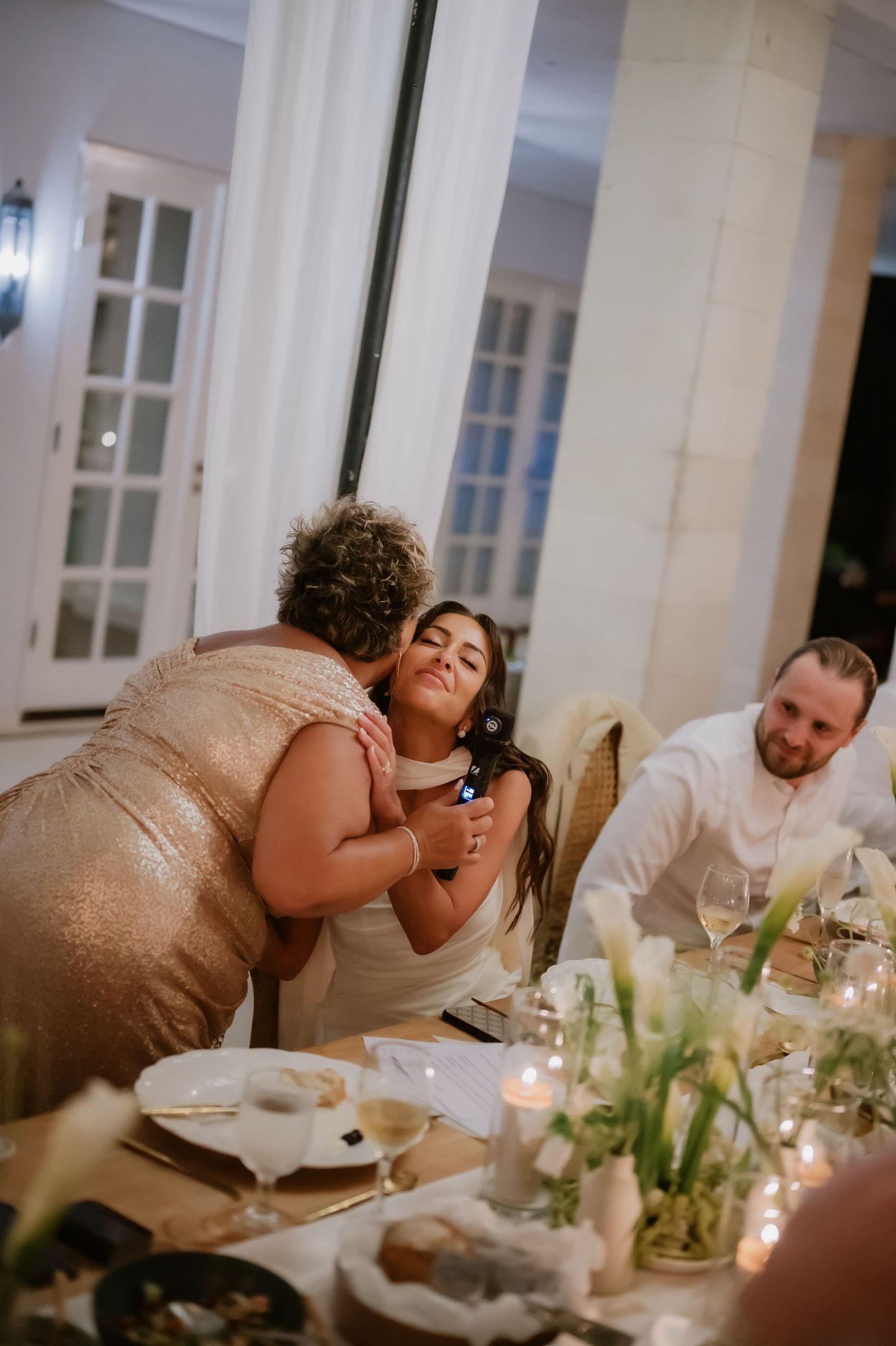 Older woman kisses bride on cheek during candlelit wedding reception dinner at the table.