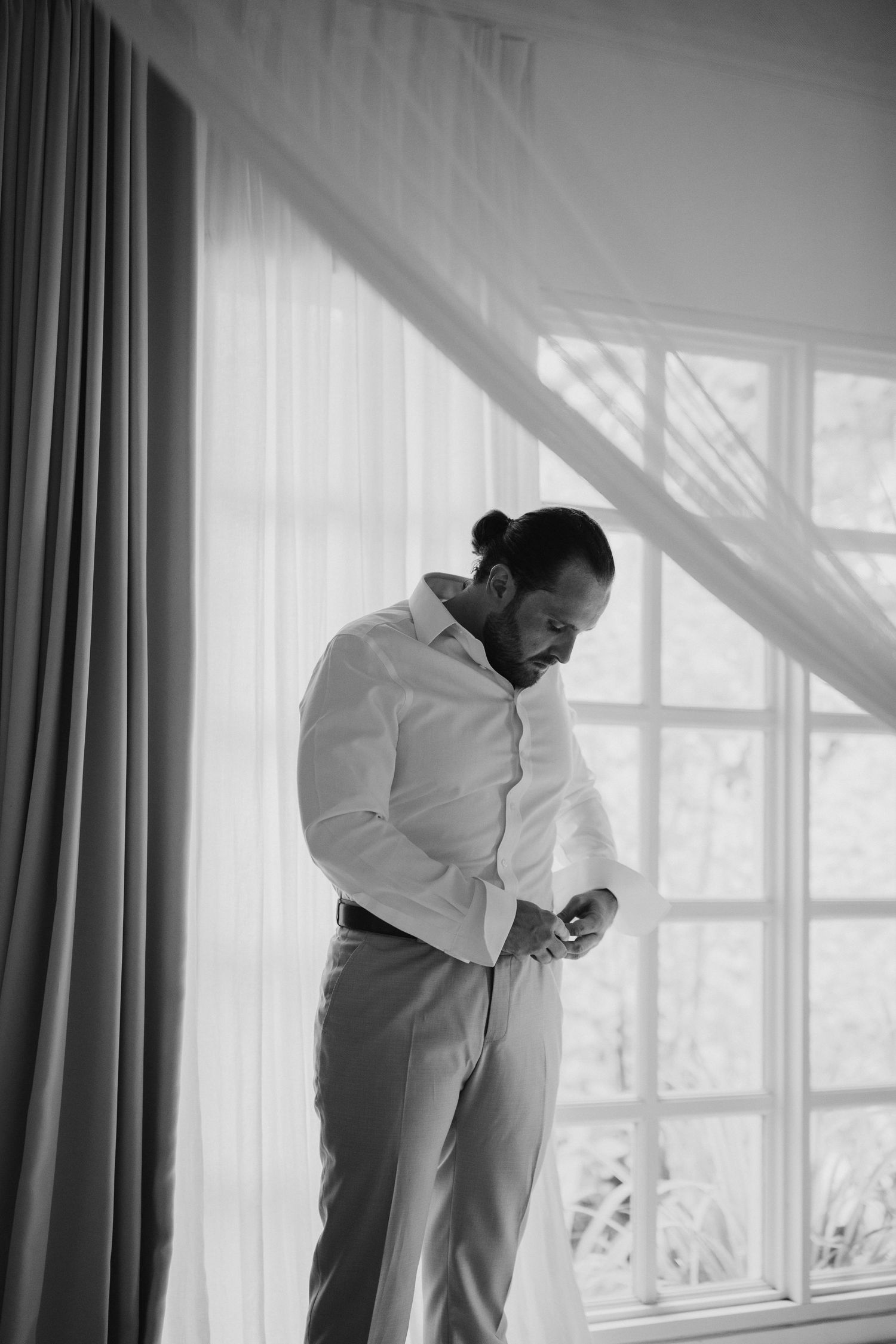 Black and white portrait of a groom buttoning his shirt by a bright window before the wedding.