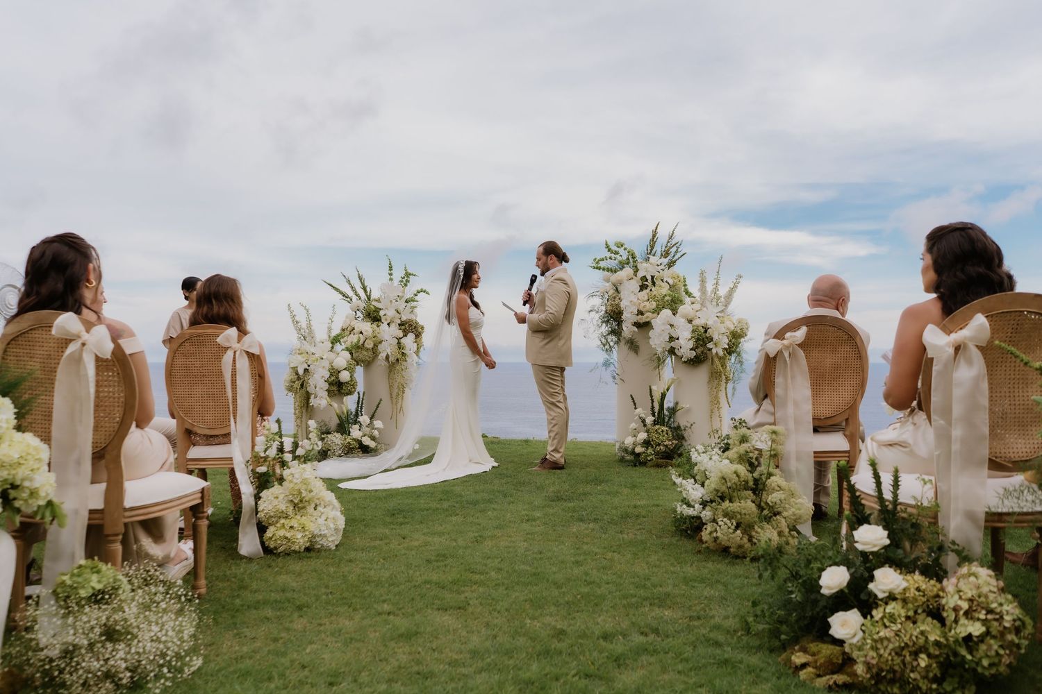 Outdoor oceanfront wedding ceremony with bride and groom at the altar surrounded by white floral arrangements.