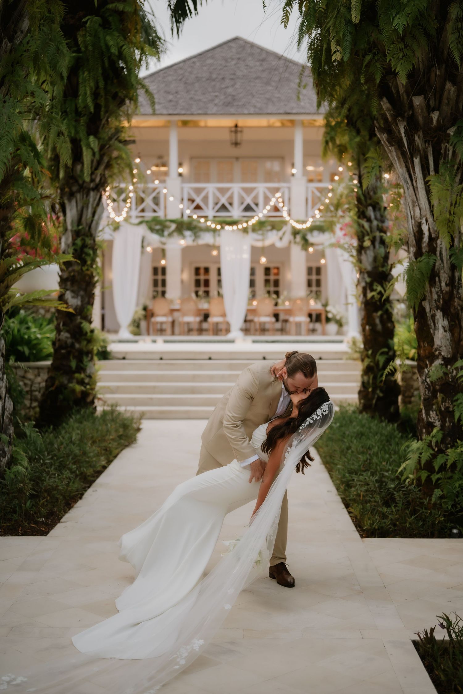 Groom dips bride in a romantic kiss on a garden path before an elegant tropical wedding venue at dusk.