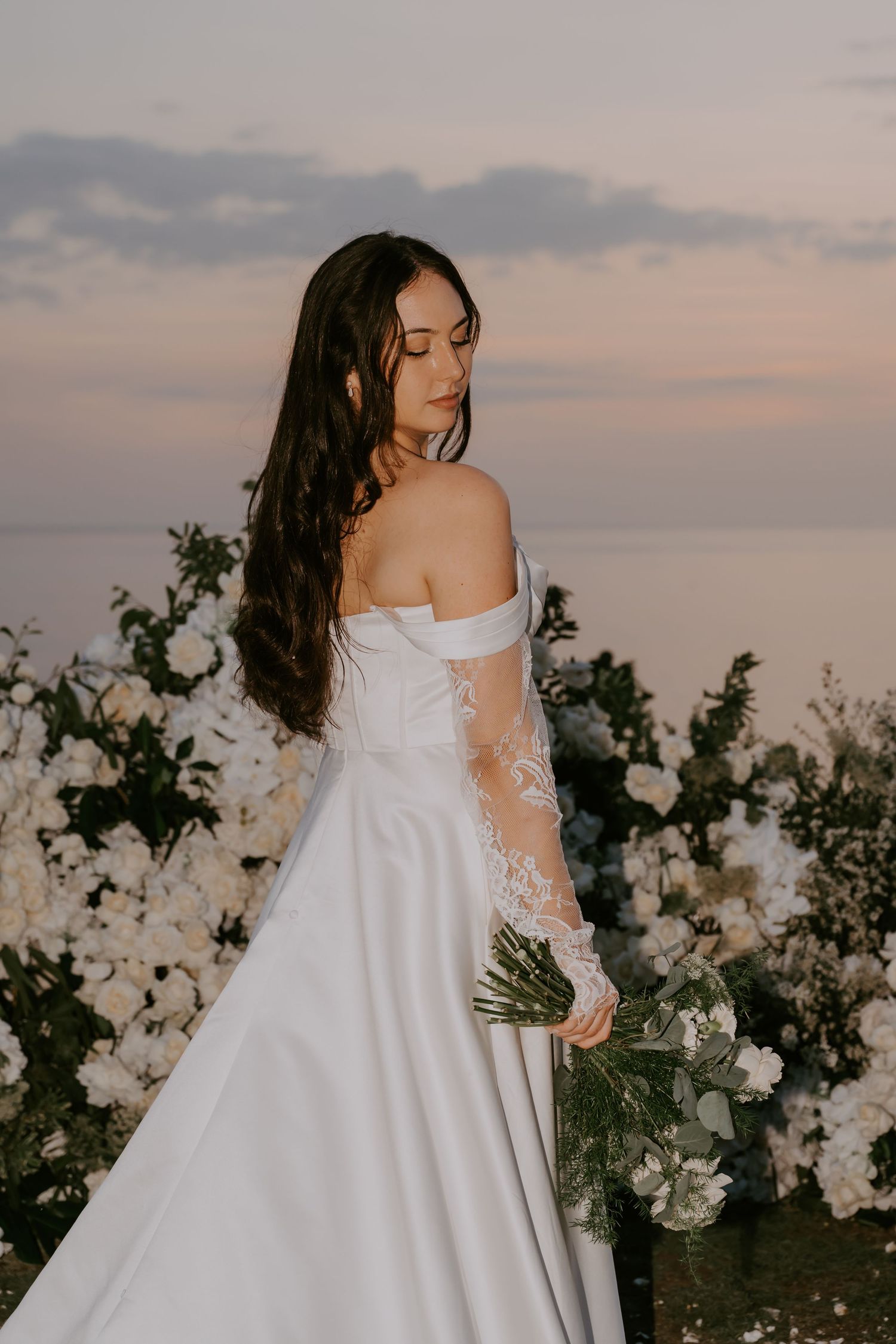 Bride in off-shoulder white satin gown with lace sleeves holds bouquet beside white floral arrangements at sunset.