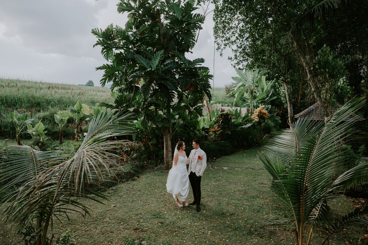 Bride and groom walk hand in hand through a lush tropical garden after their wedding ceremony.