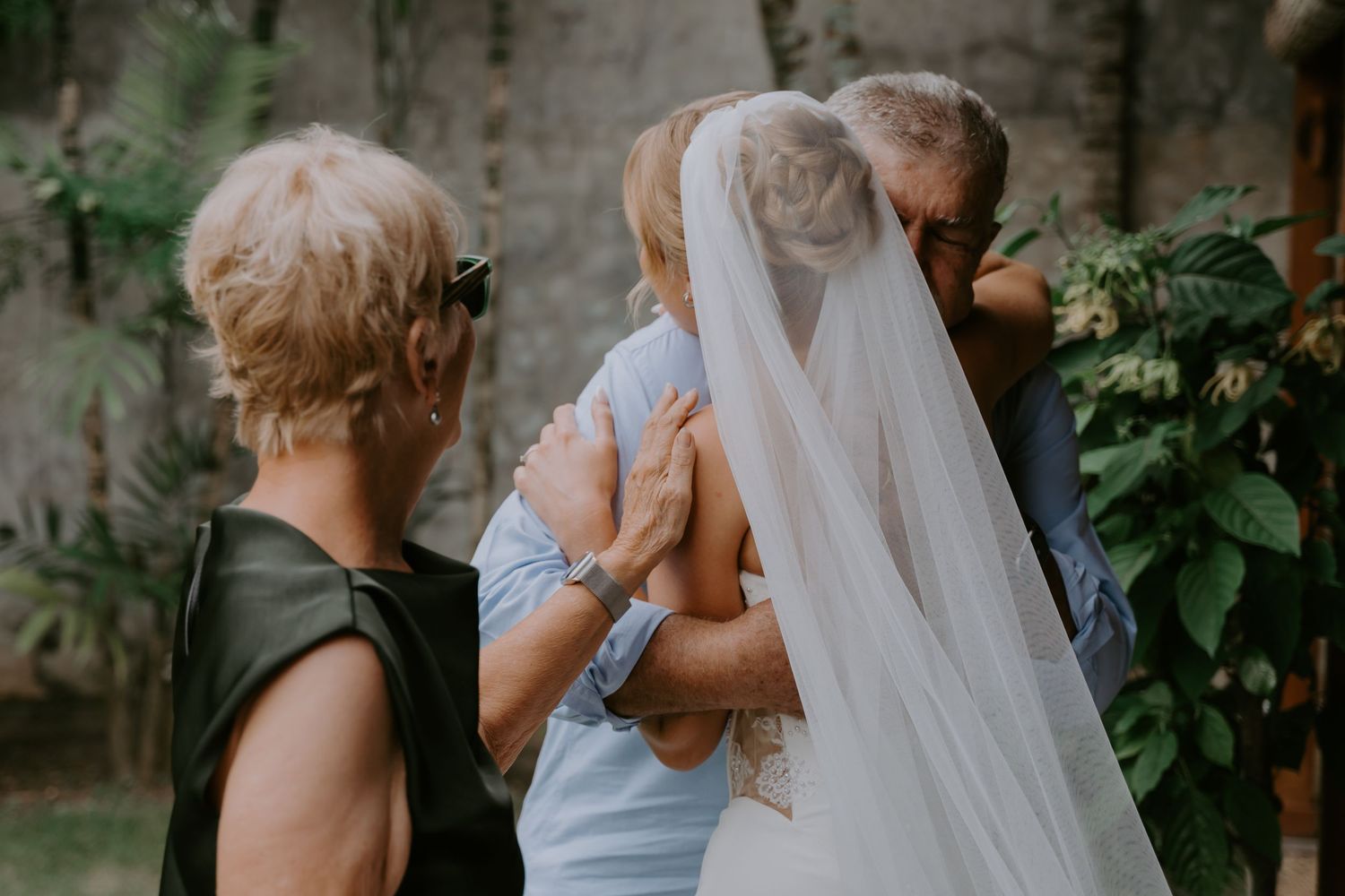 Bride in white veil embraced by elderly family members during an outdoor wedding ceremony.