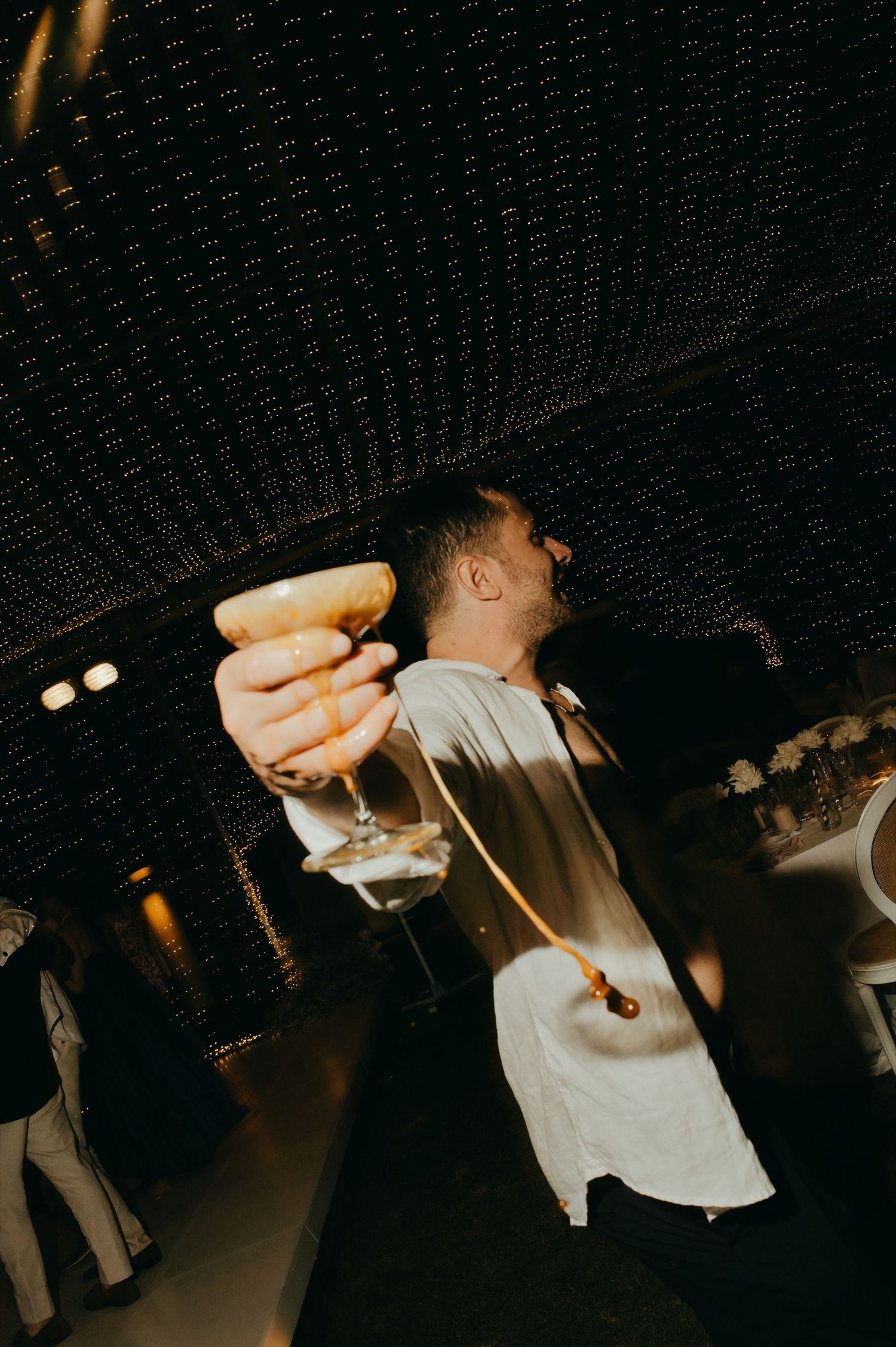 A man raises a cocktail glass triumphantly at an elegant evening event under a woven ceiling.
