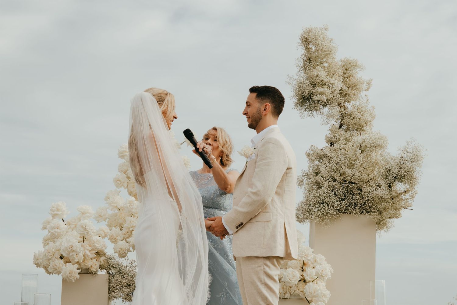 Bride and groom exchanging vows at outdoor altar surrounded by white floral arrangements.