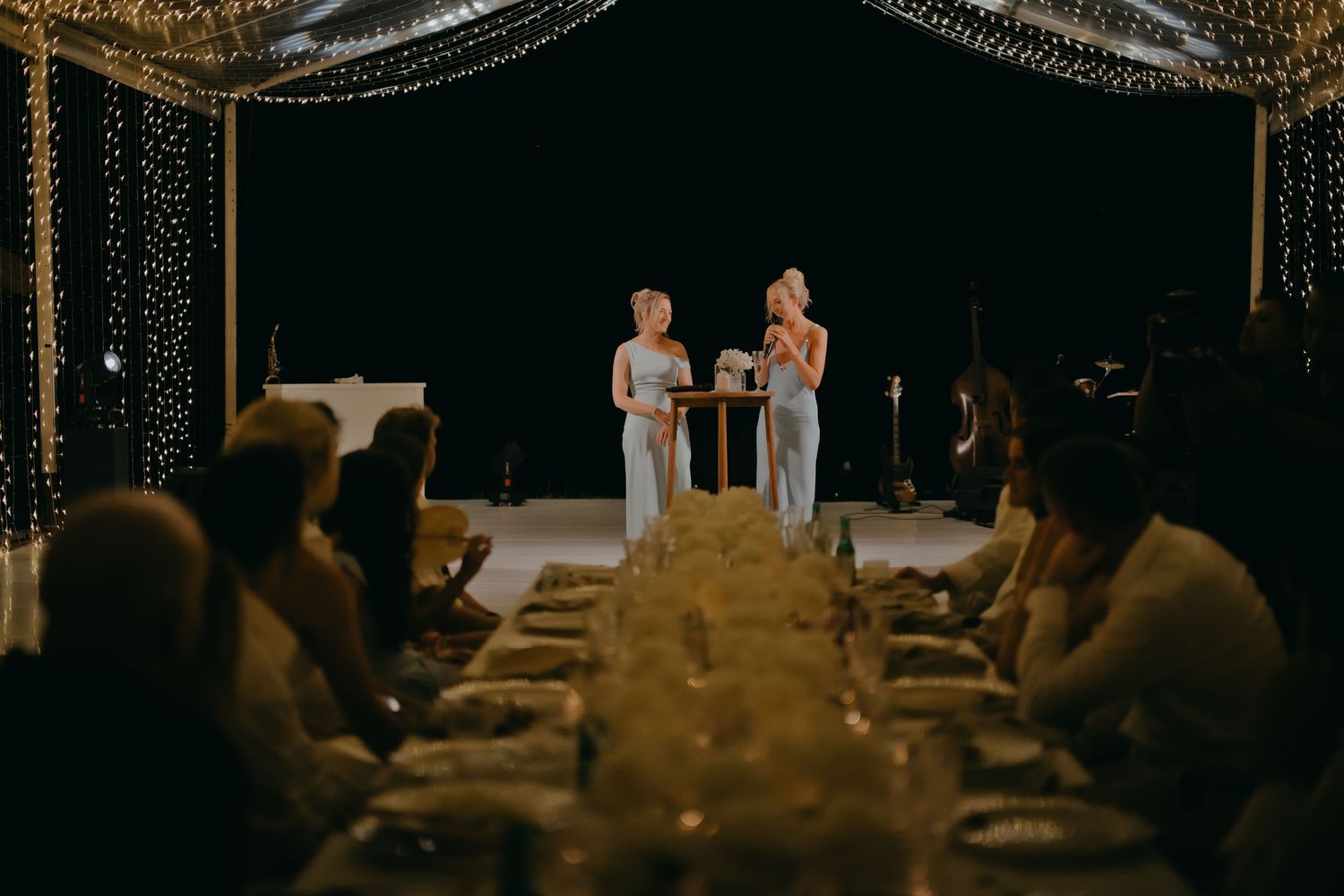 Two bridesmaids in blue dresses give speeches at candlelit wedding reception dinner table.