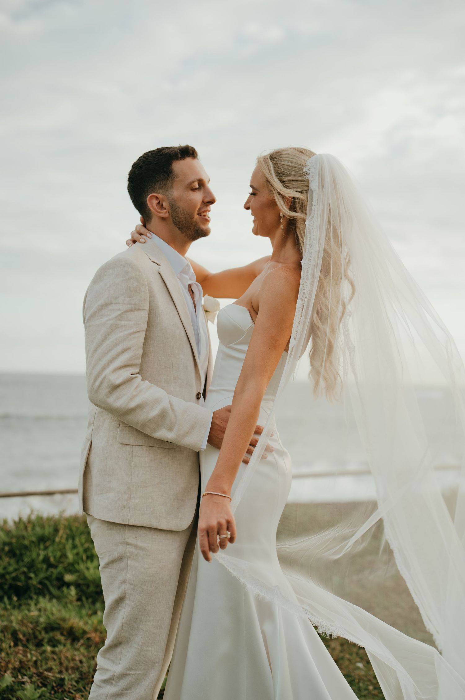 Bride and groom share a romantic moment by the ocean, veil flowing in the breeze.