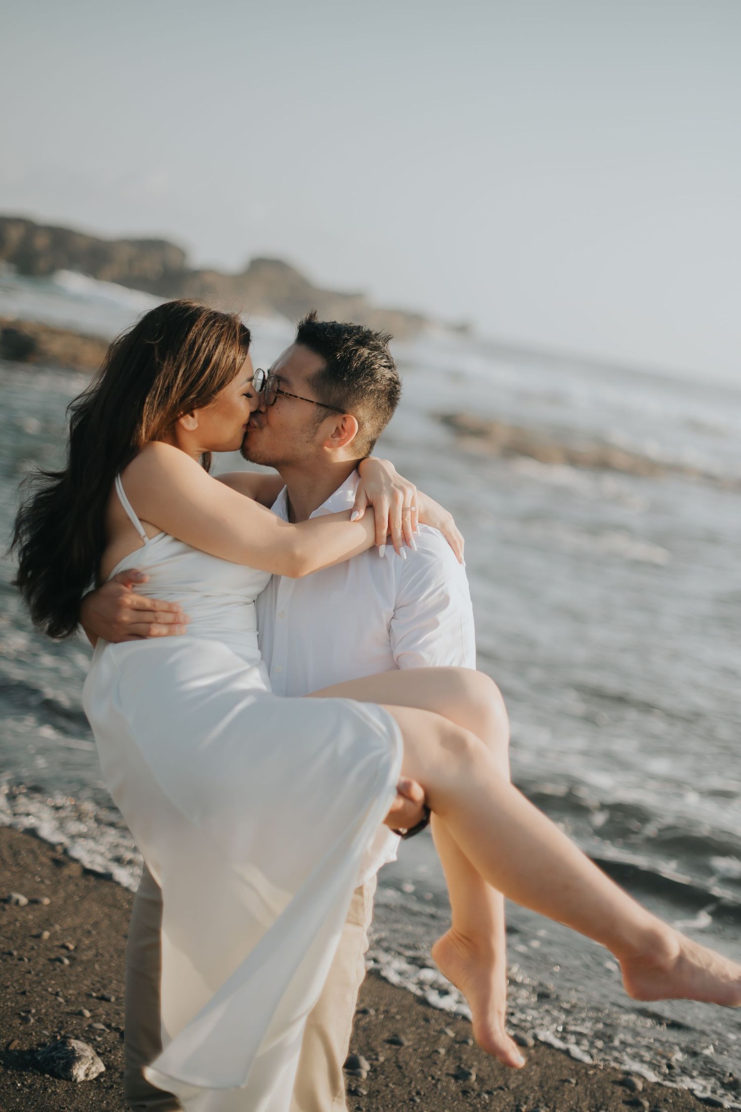 A couple shares a romantic kiss on a rocky beach, with the groom carrying his bride in a white dress by the ocean.