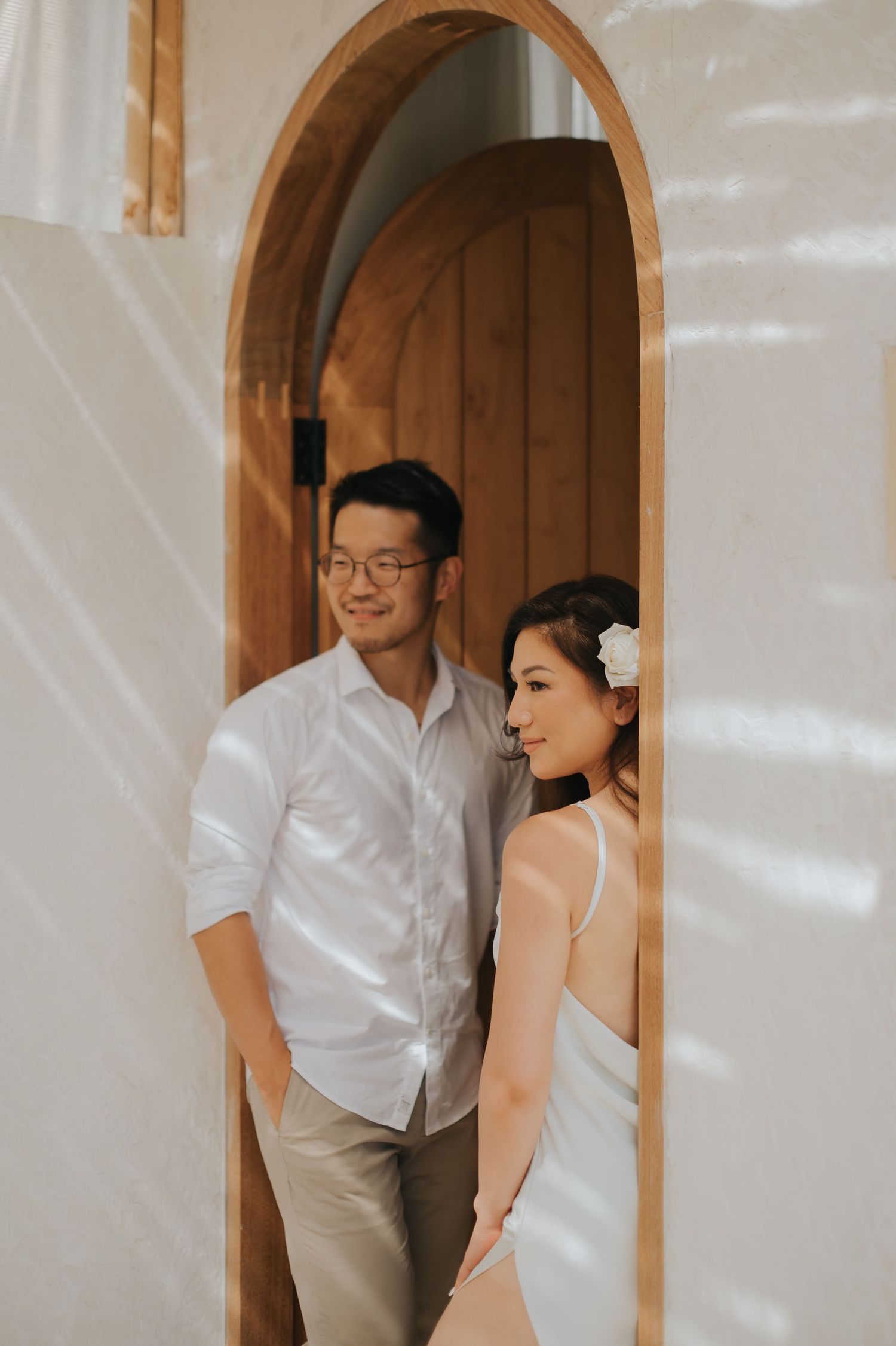 A couple in white attire poses in an arched wooden doorway, the woman wearing a floral hair accessory.