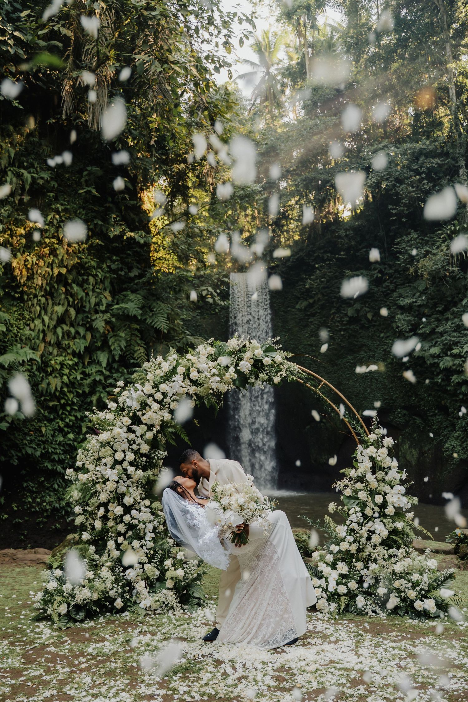 Couple sharing first kiss under white floral arch with waterfall backdrop and falling flower petals.