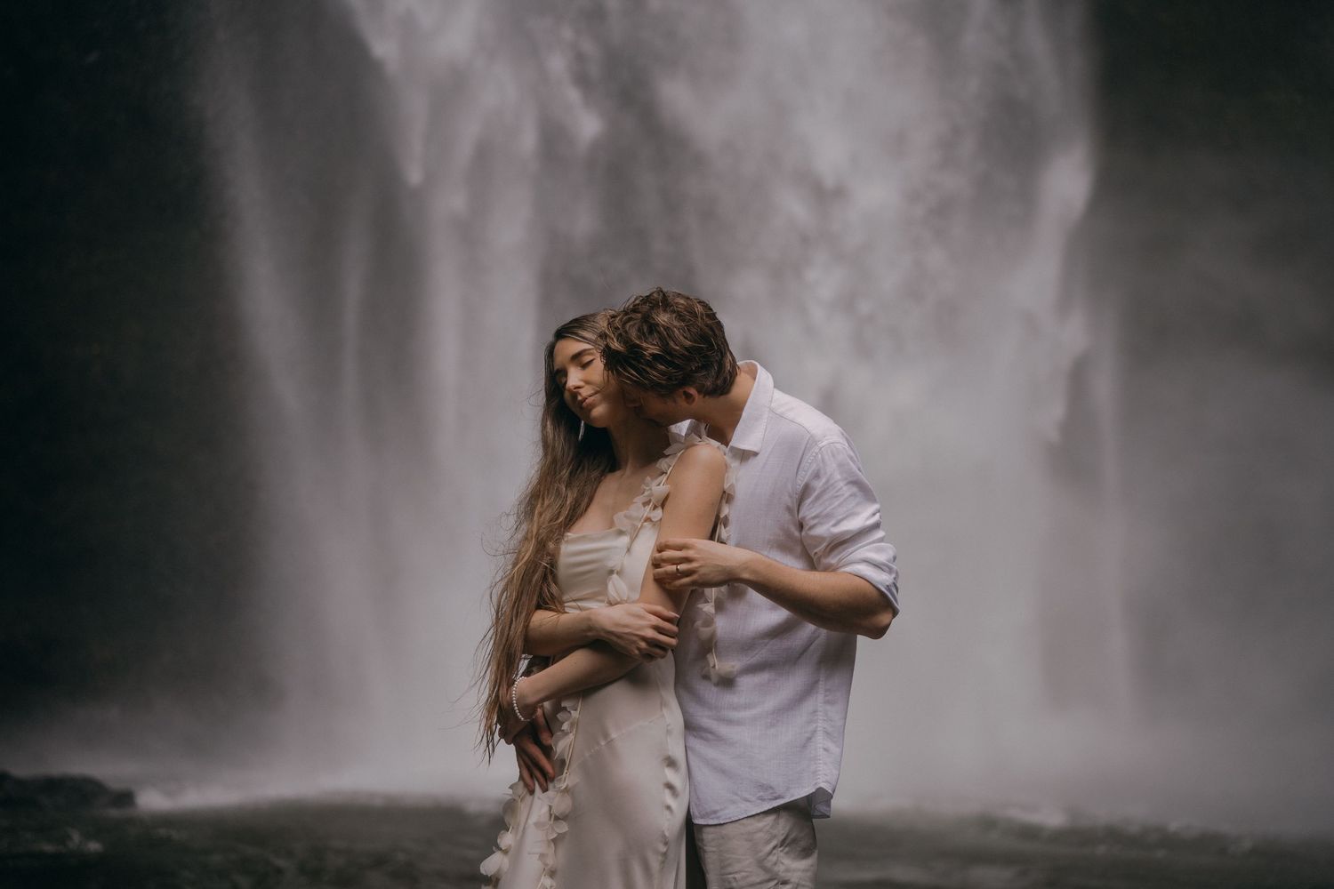 A couple embraces in front of a misty waterfall backdrop creating a romantic silhouette against the cascading water.
