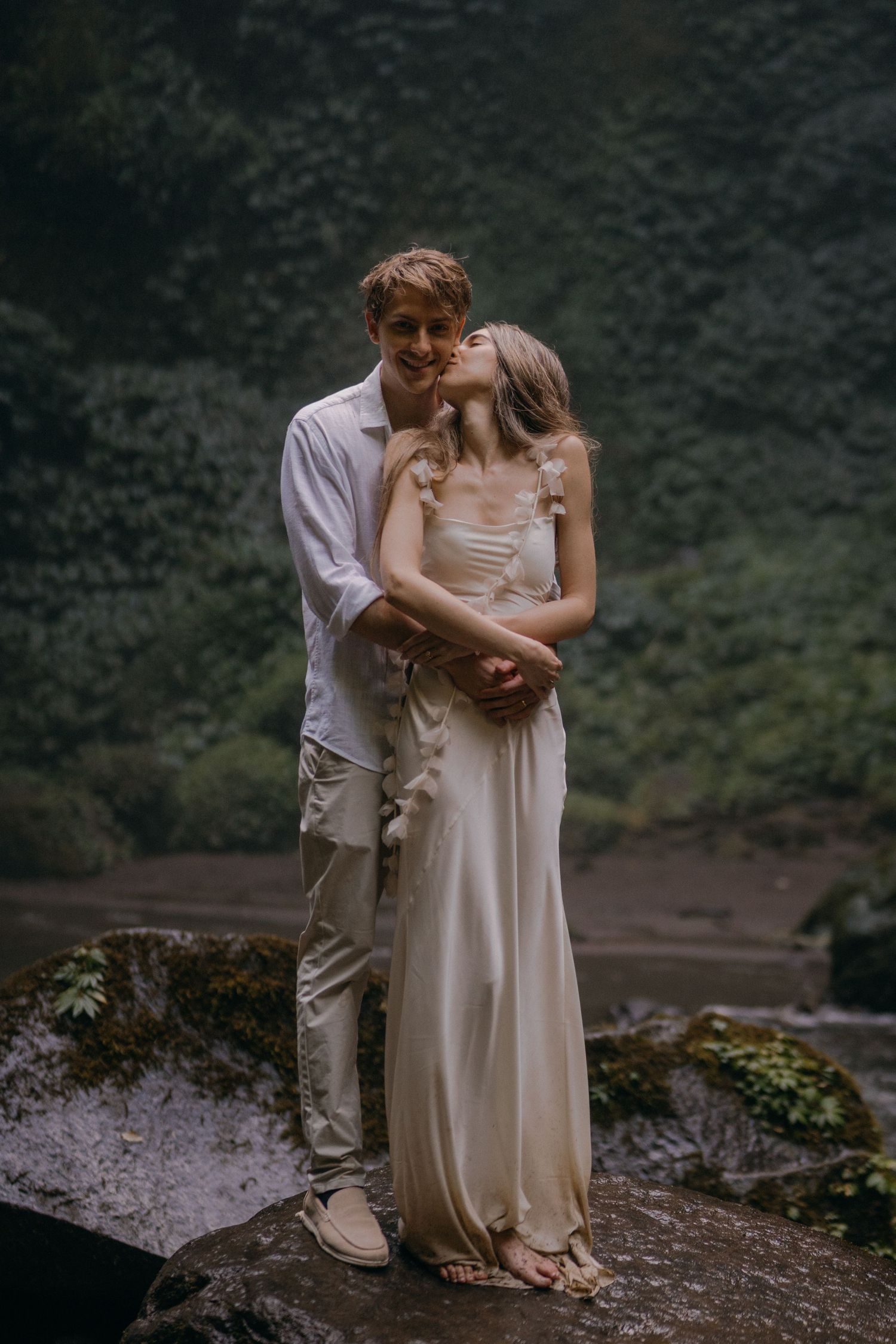 A couple in wedding attire shares an intimate moment on a mountainside overlooking a scenic valley landscape.