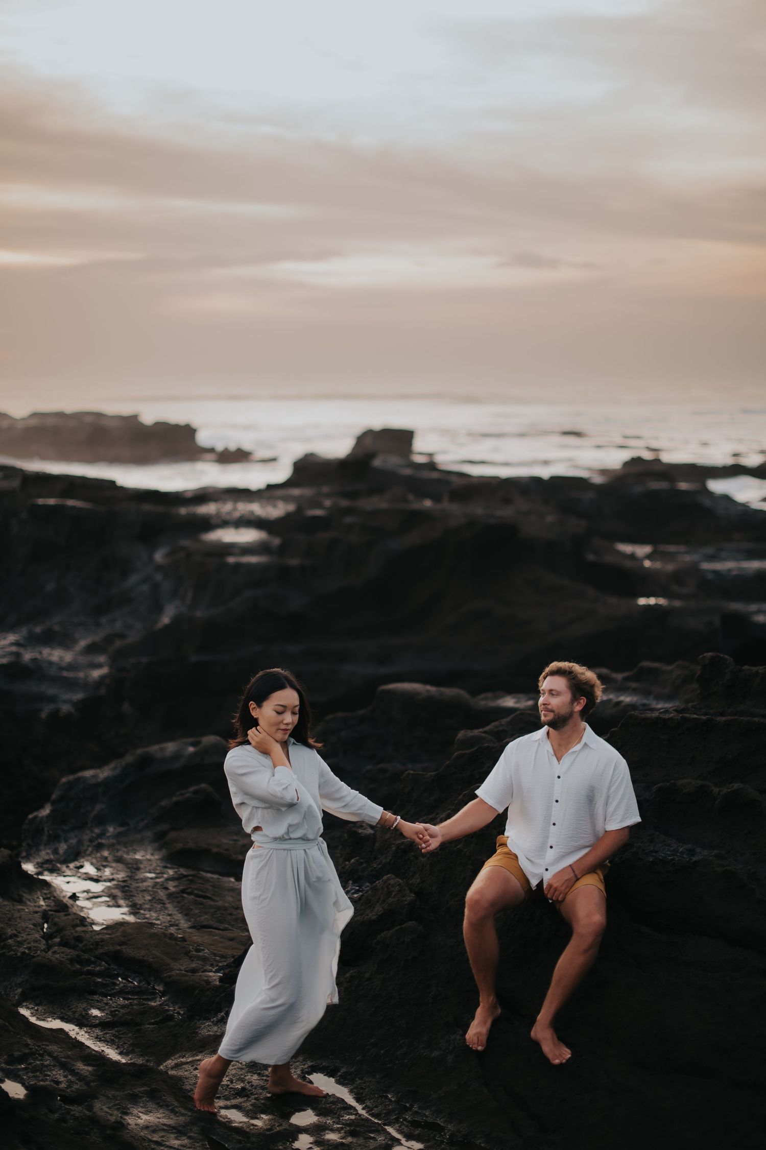 Two people dance and hold hands on dark rocky cliffs at sunset by the ocean.