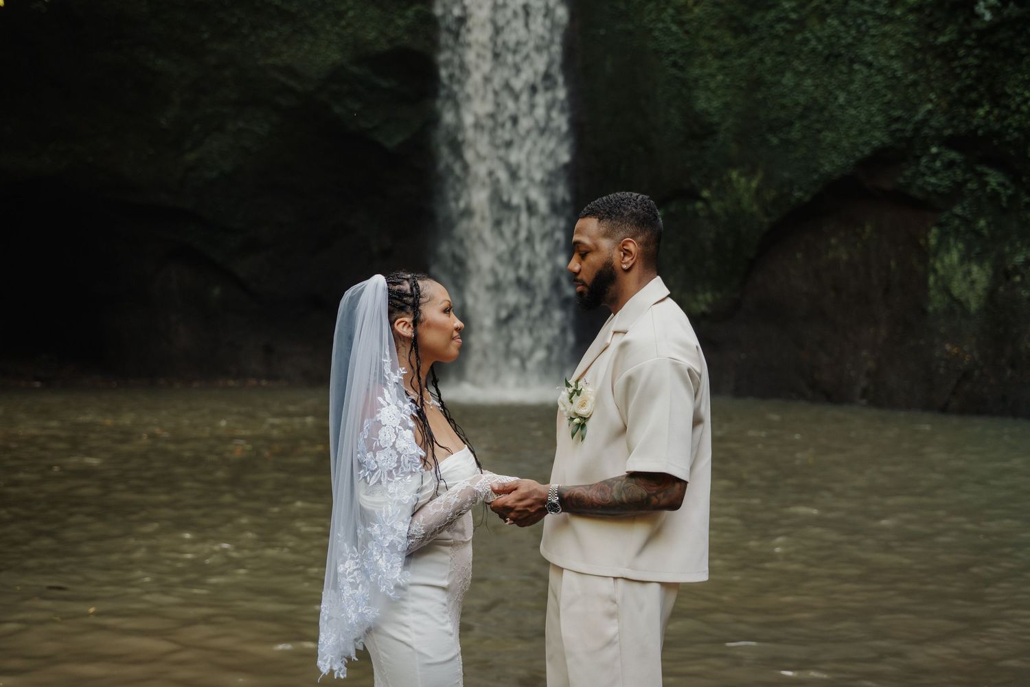Bride and groom hold hands facing each other standing in water before a stunning tropical waterfall backdrop.