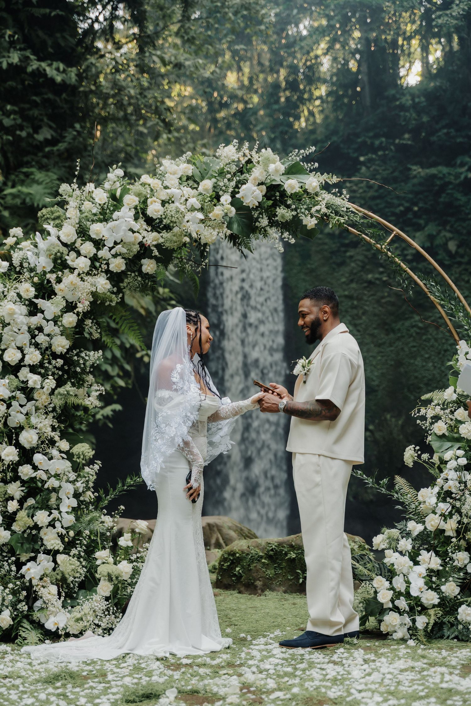 A couple exchanges wedding rings beneath a circular white floral arch with a waterfall behind them.