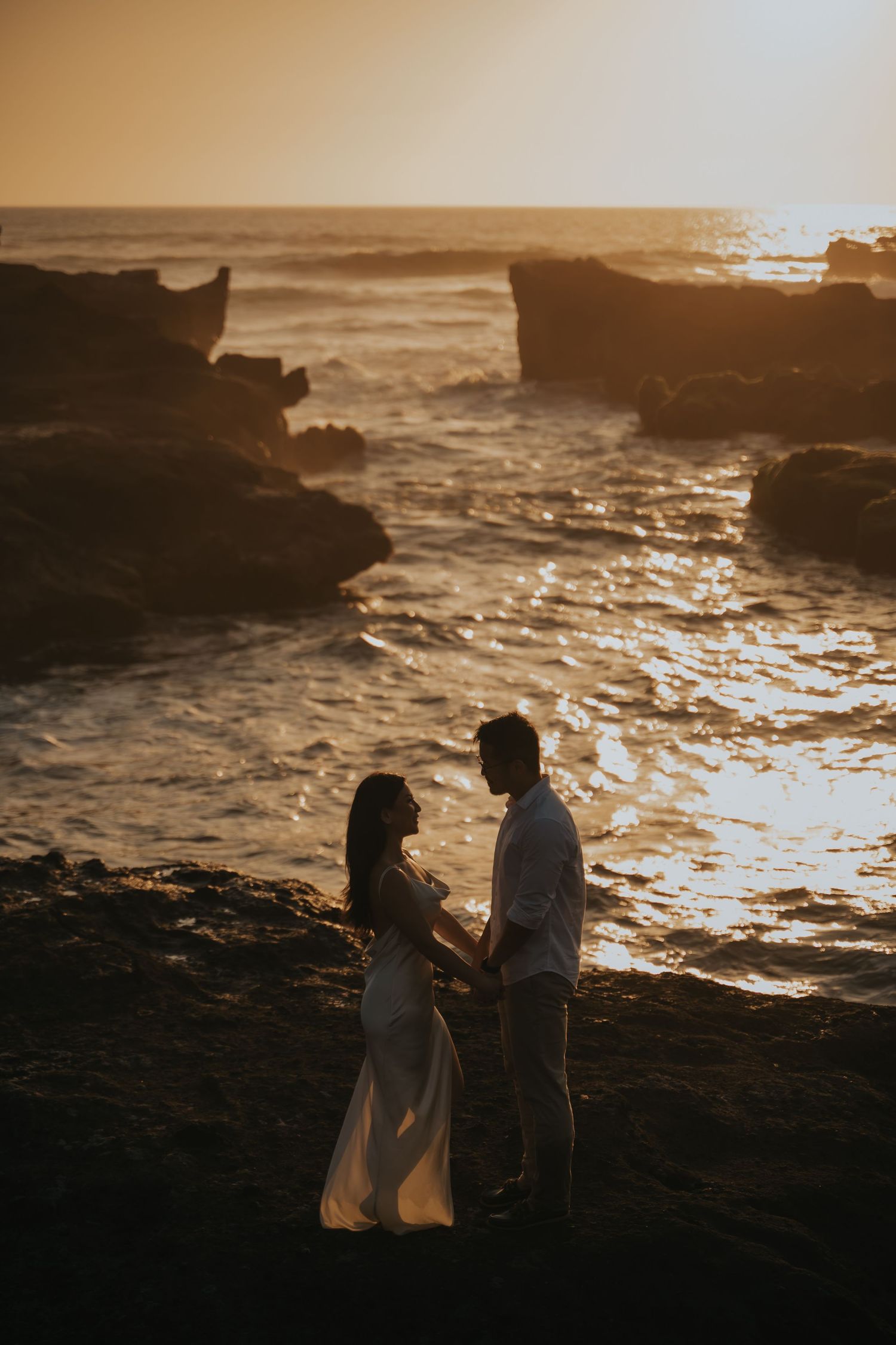 Couple holding hands at sunset on rocky coastline, silhouetted against golden ocean waves.