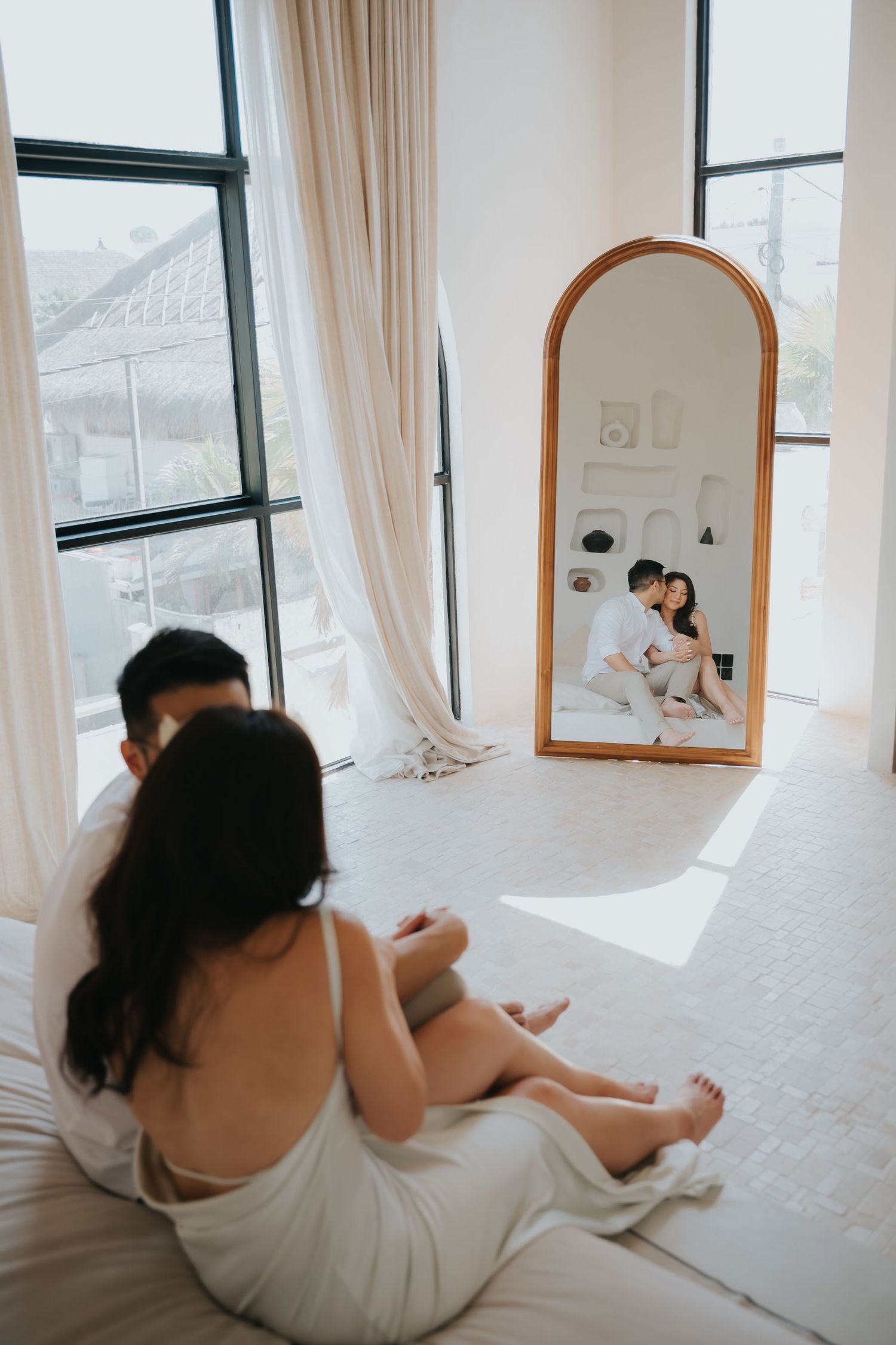 Couple sitting on a white bed, reflected in an arched mirror, in a bright minimalist bedroom with sheer curtains.