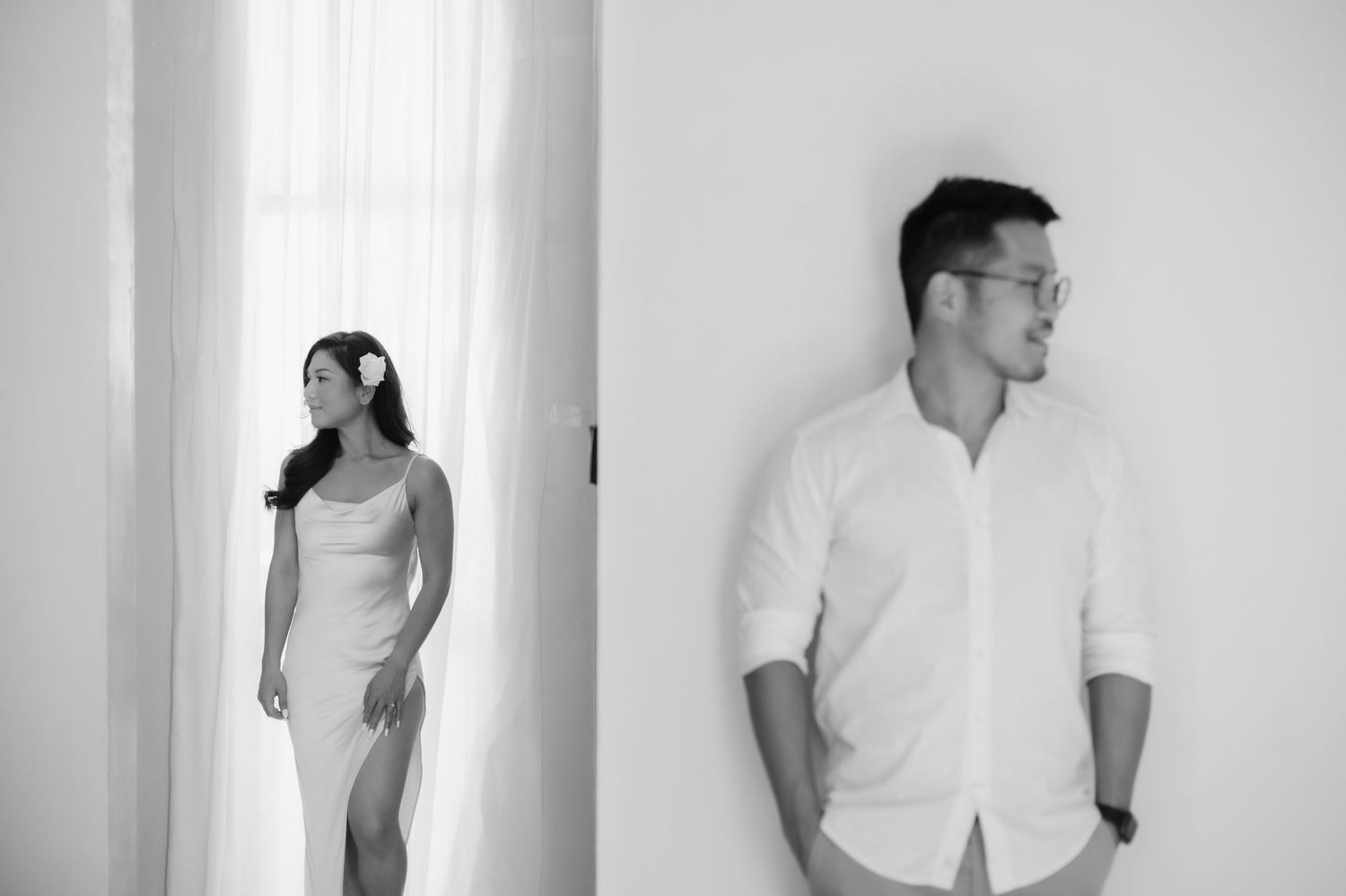 Black and white photo of a couple, woman in white dress by curtains, man in white shirt against wall.