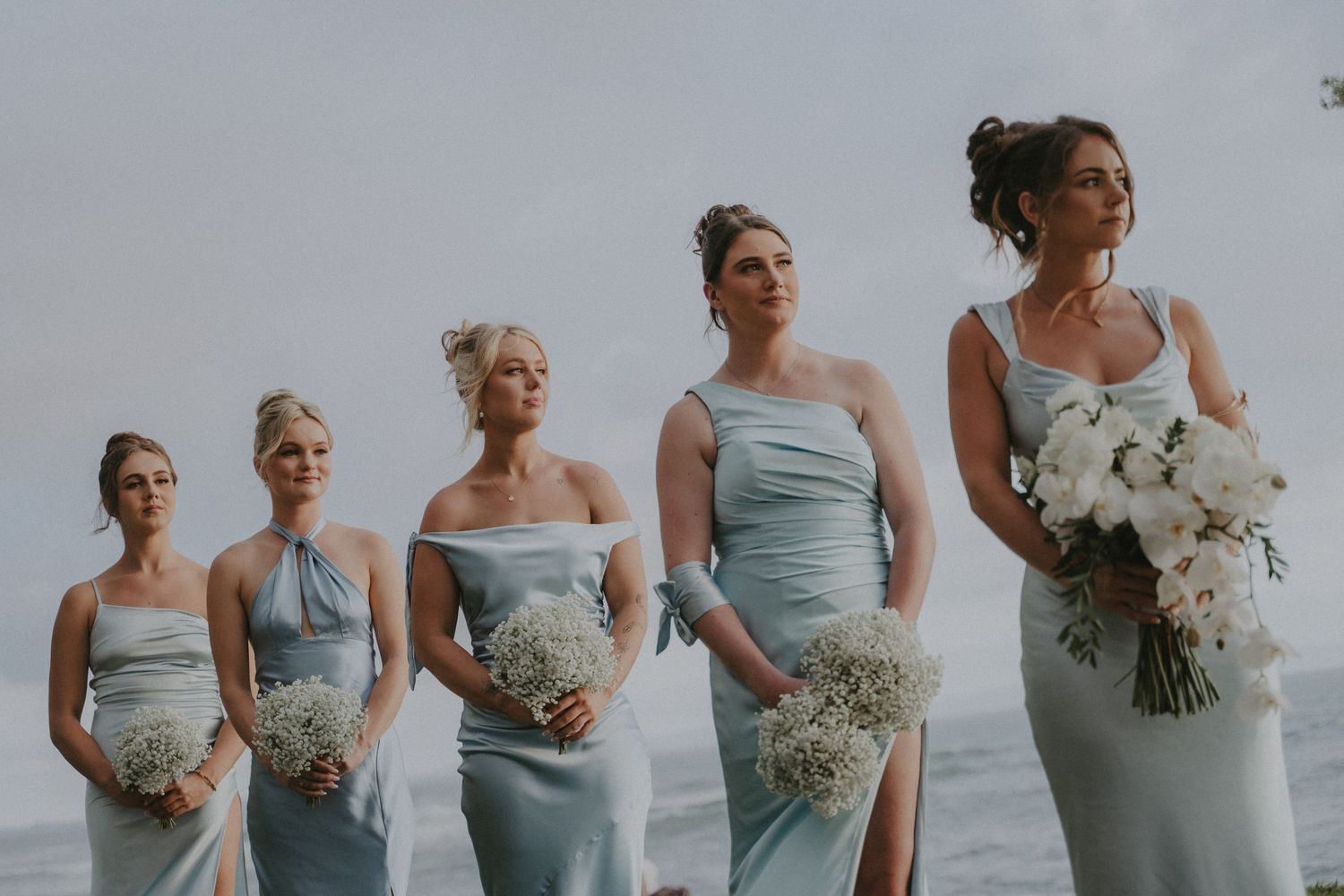 Bridesmaids in light blue dresses holding white bouquets pose together for wedding photos on a cloudy day.