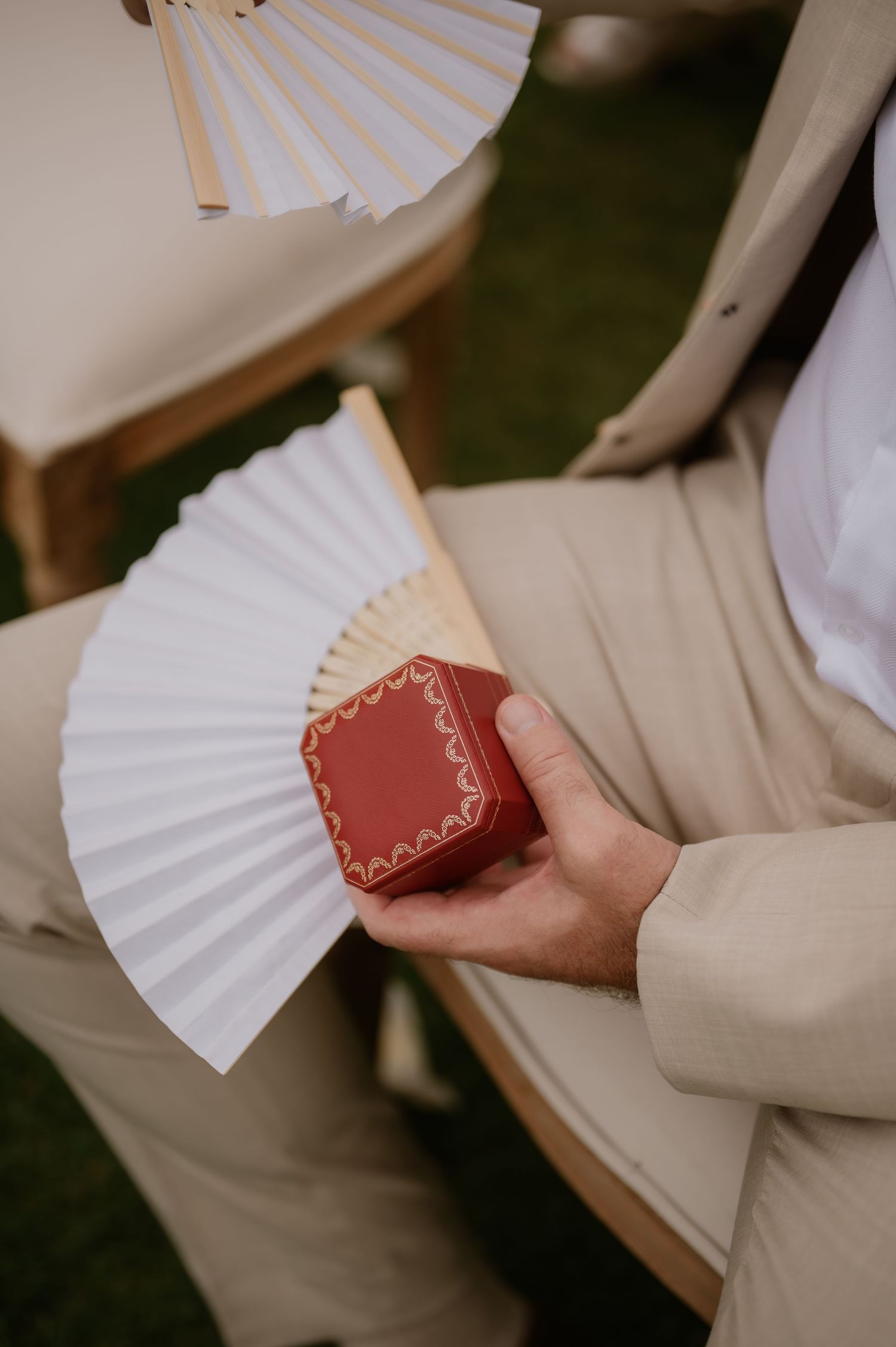 Guest holding a red Cartier ring box with white paper fans at an outdoor wedding ceremony.