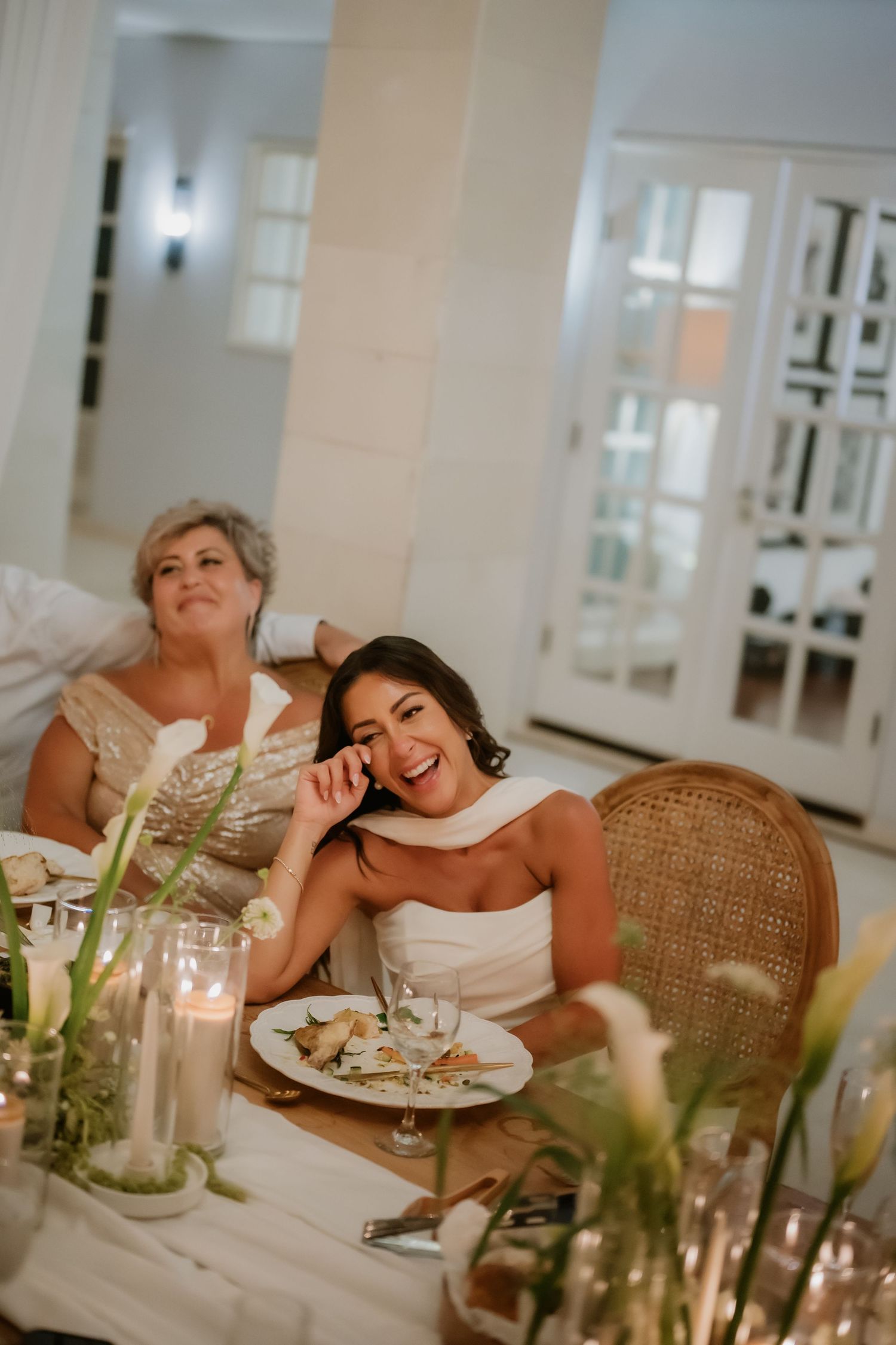 Bride laughs at candlelit wedding reception table seated beside woman in gold sequin dress.