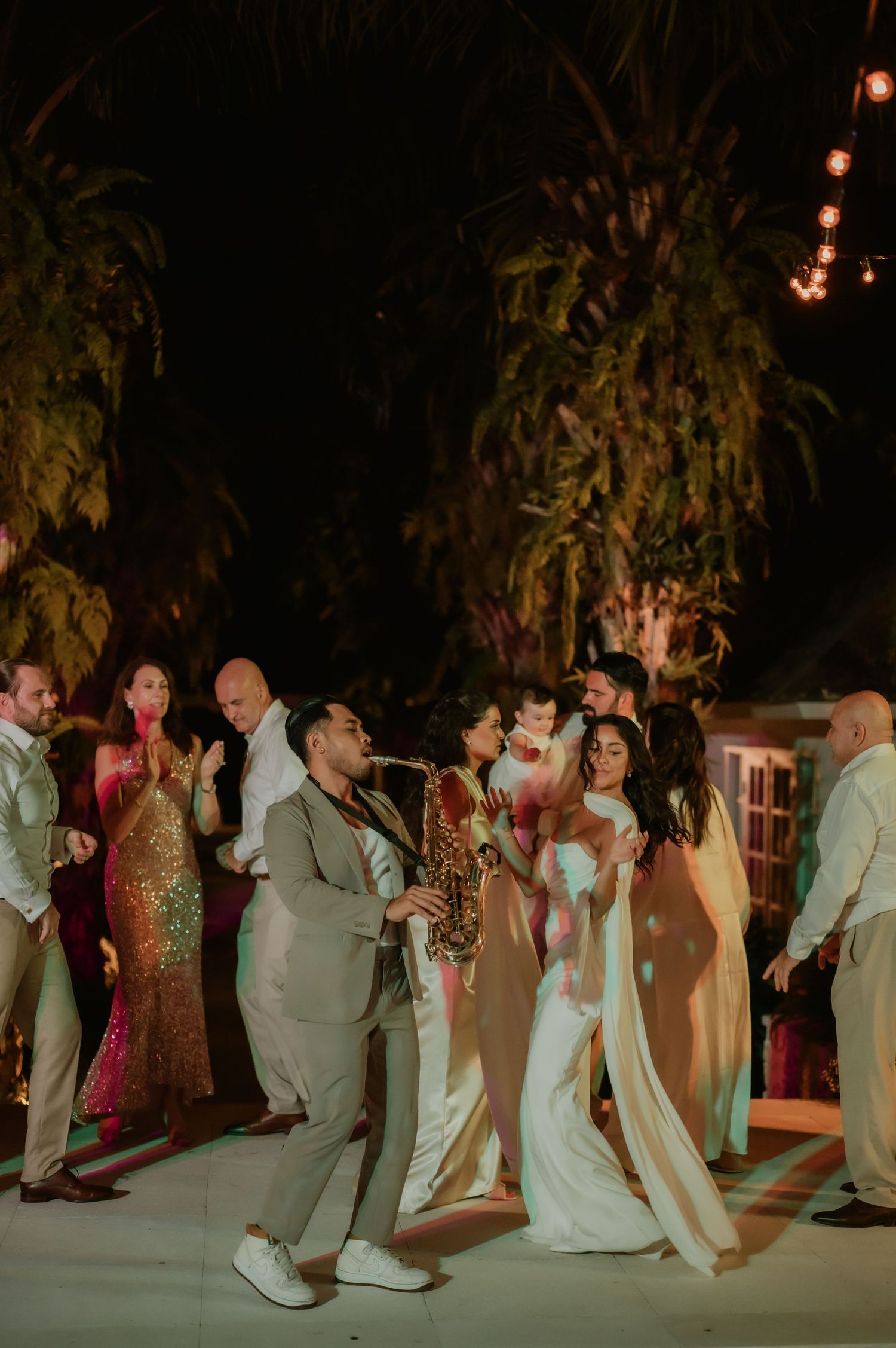 Bride groom and guests dance joyfully outdoors under string lights at lively wedding reception.