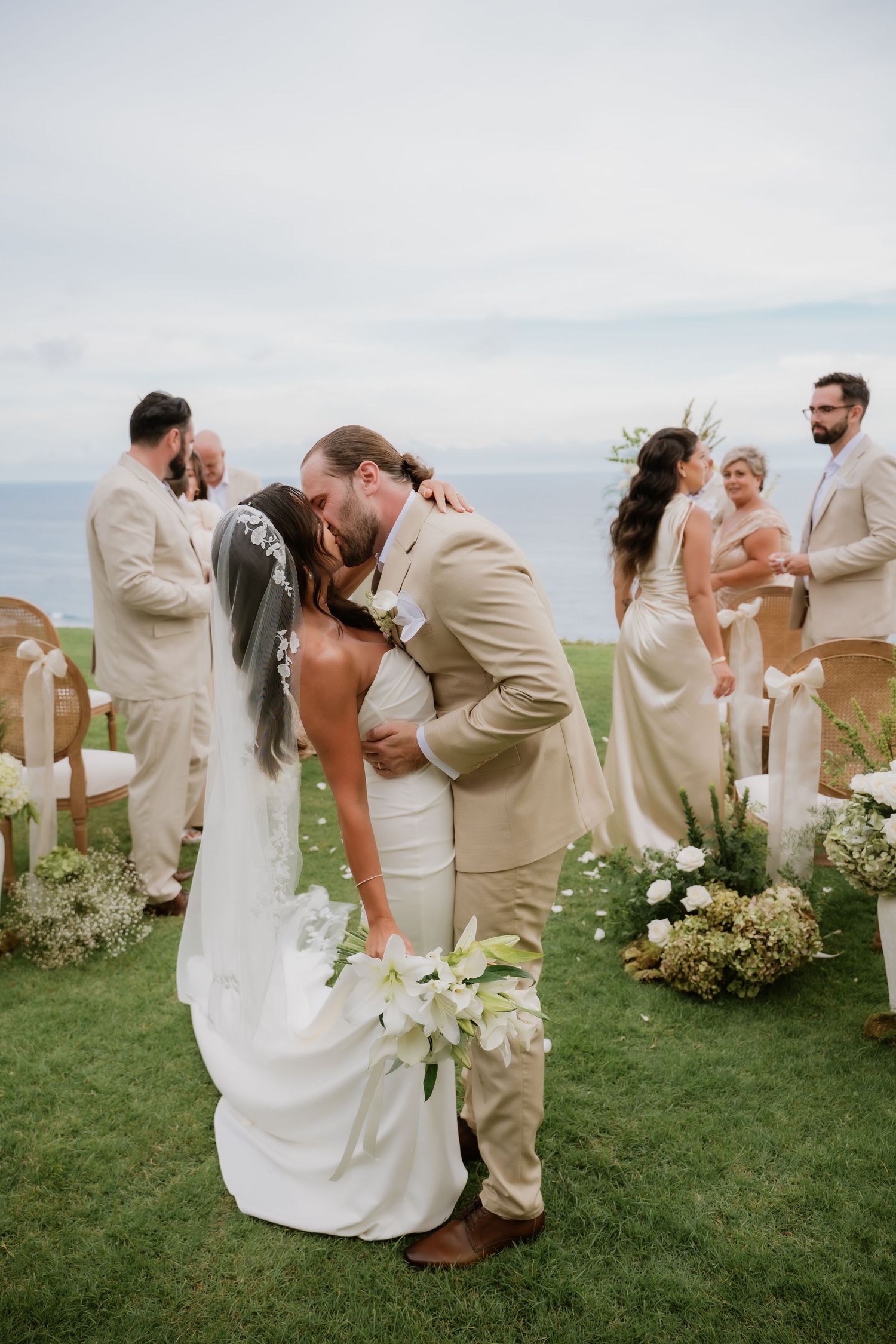 Newlyweds share first kiss on oceanfront lawn surrounded by guests in neutral attire after ceremony.