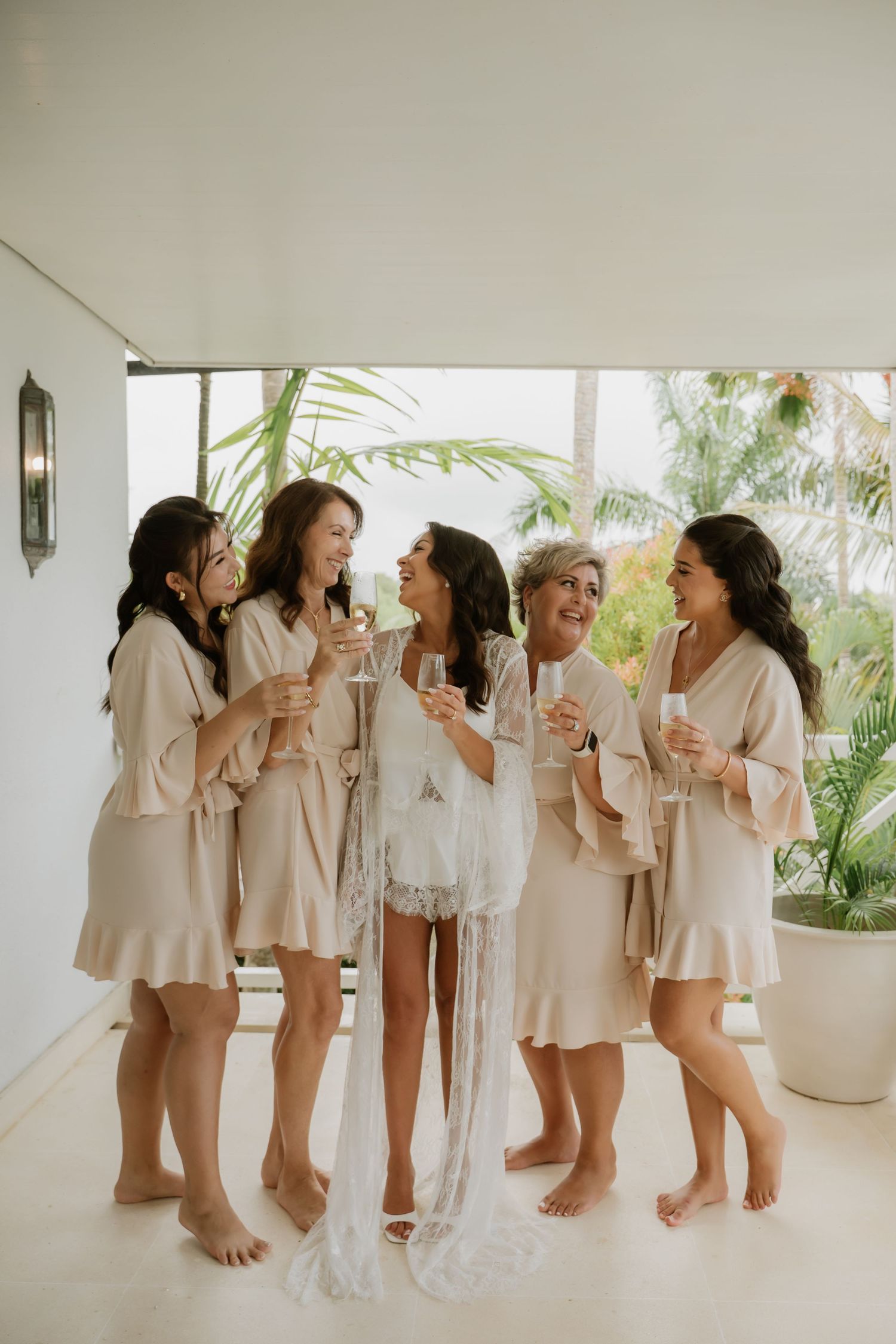 Five bridesmaids in matching beige robes toasting with champagne glasses before a tropical wedding.
