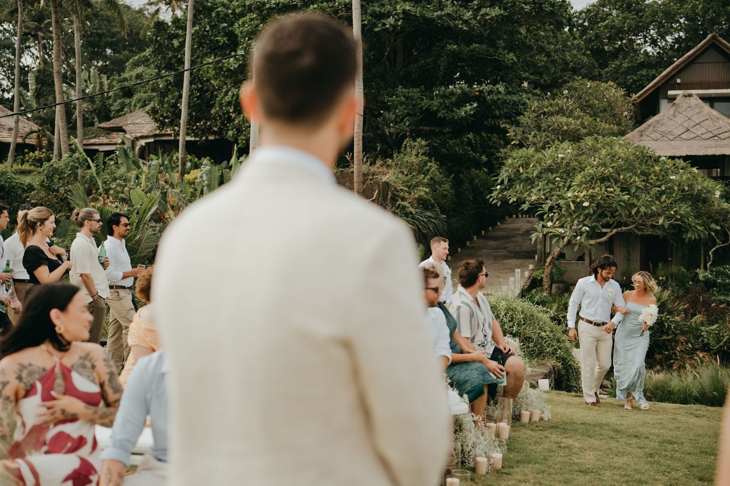 Groom stands at outdoor wedding ceremony aisle as guests are seated in lush tropical garden setting.
