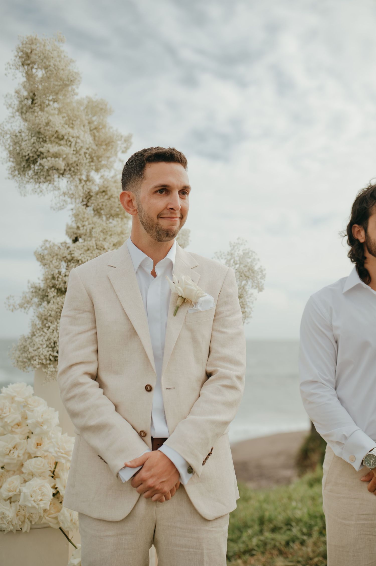 Groom in beige linen suit with white boutonniere stands at outdoor wedding ceremony altar.