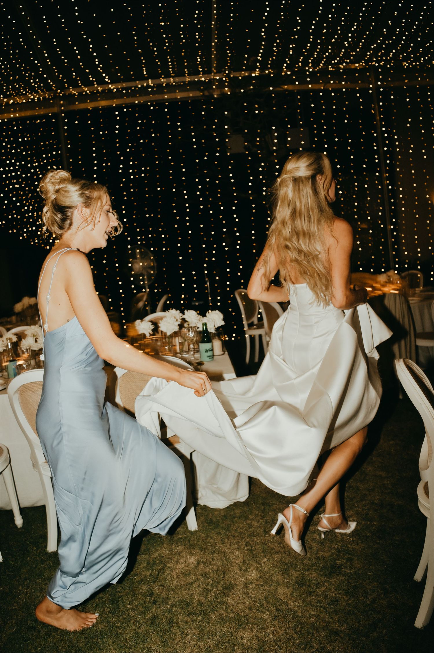 Bridesmaid in blue gown helps bride with white wedding dress under fairy light canopy at reception.