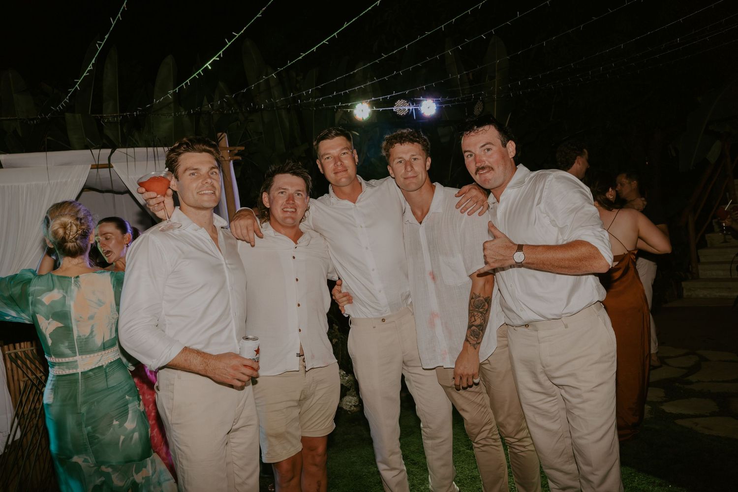 Groom poses with groomsmen in white shirts at outdoor evening wedding reception.