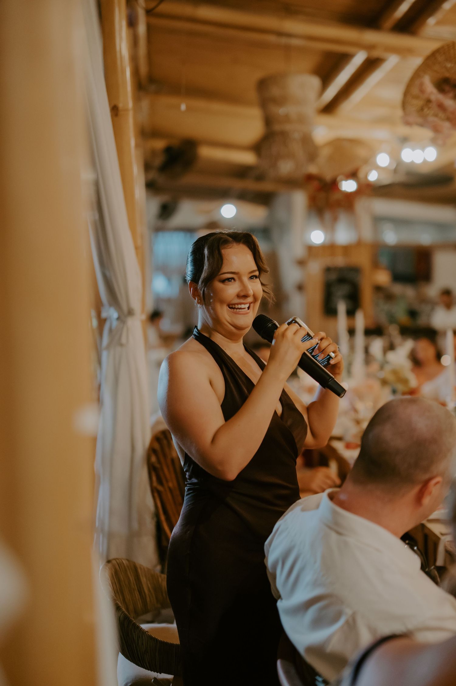 Bridesmaid in black dress delivers speech with microphone at wedding reception.