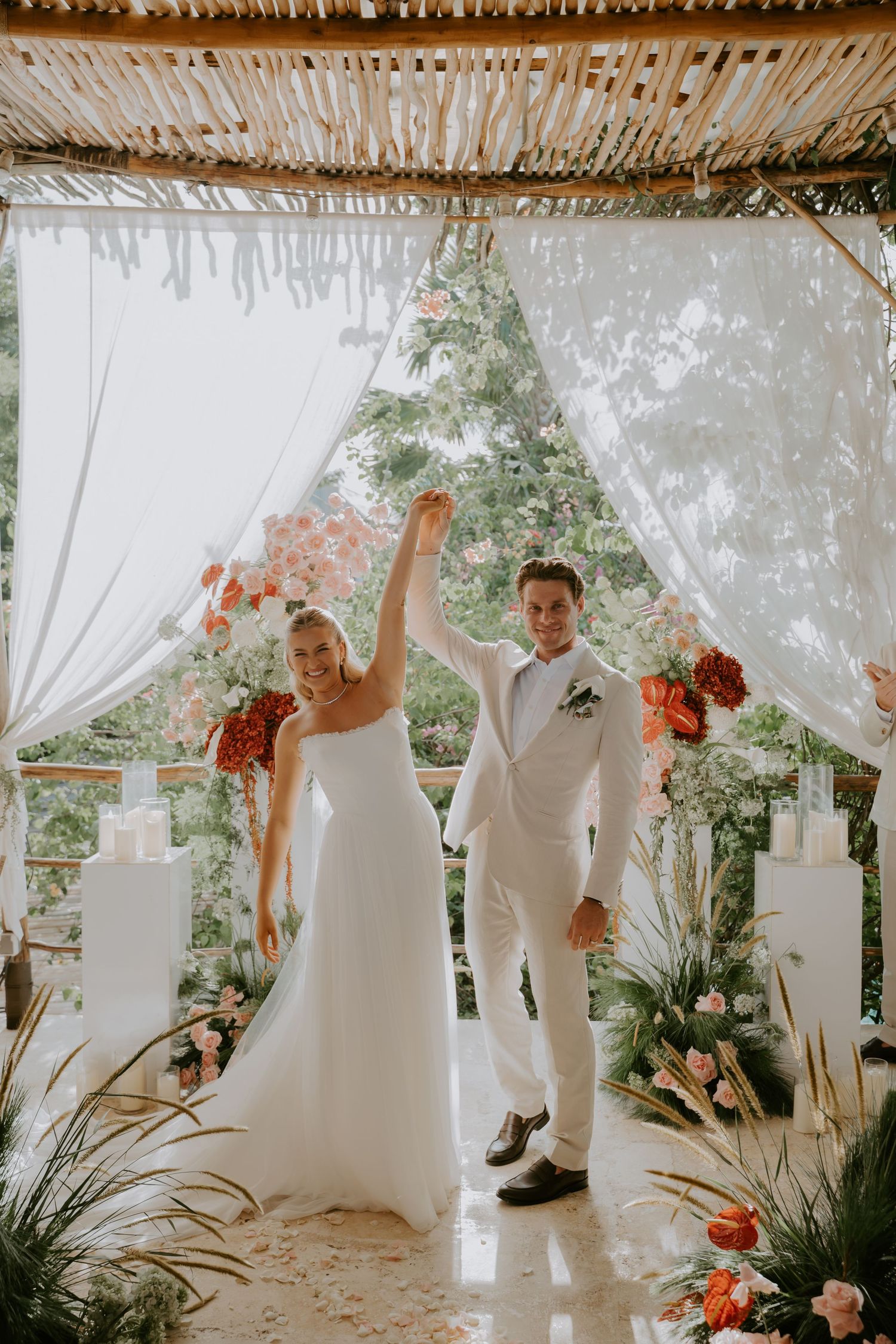 Joyful newlyweds raise their hands in celebration under a floral arch with white drapes at an outdoor wedding ceremony.