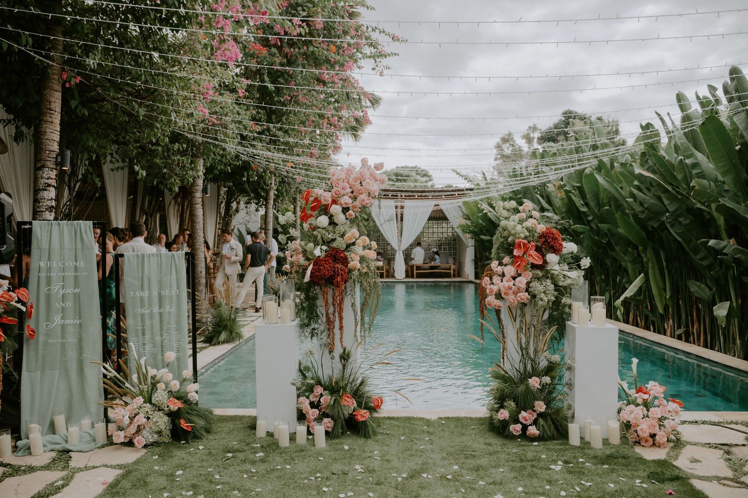 Elegant outdoor wedding entrance with floral pillars, pink and red blooms, and pool backdrop at tropical venue.