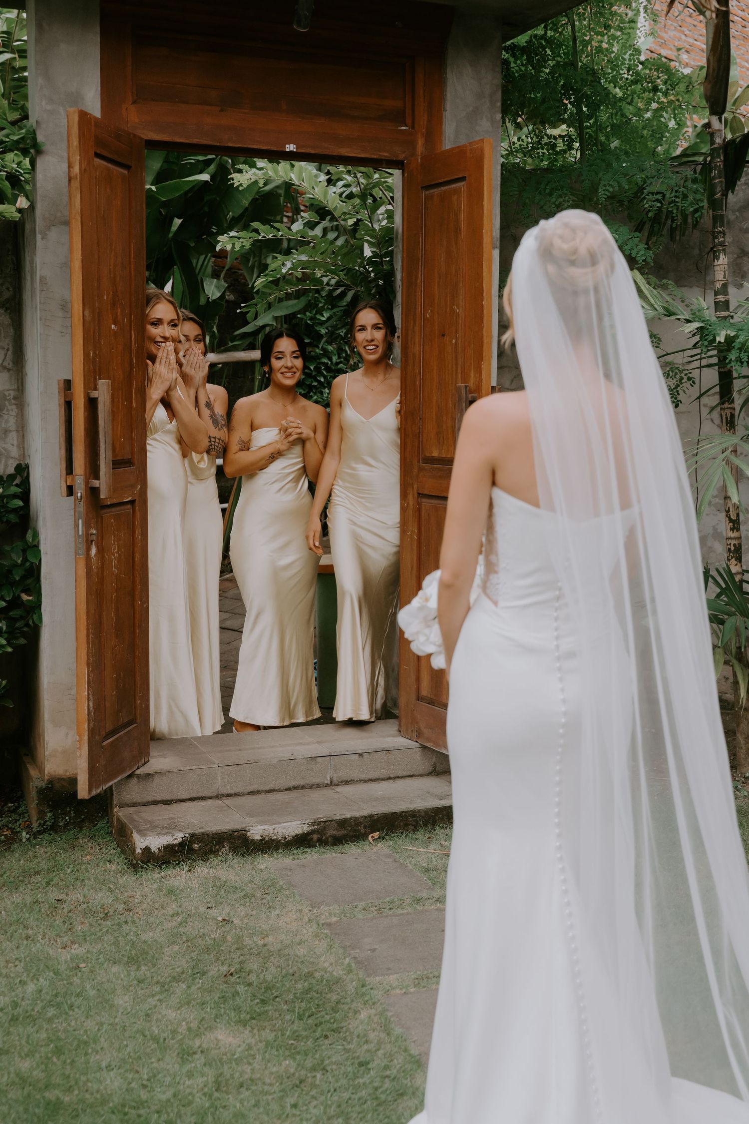 Bridesmaids in ivory satin dresses smile as bride in fitted white gown with veil approaches.