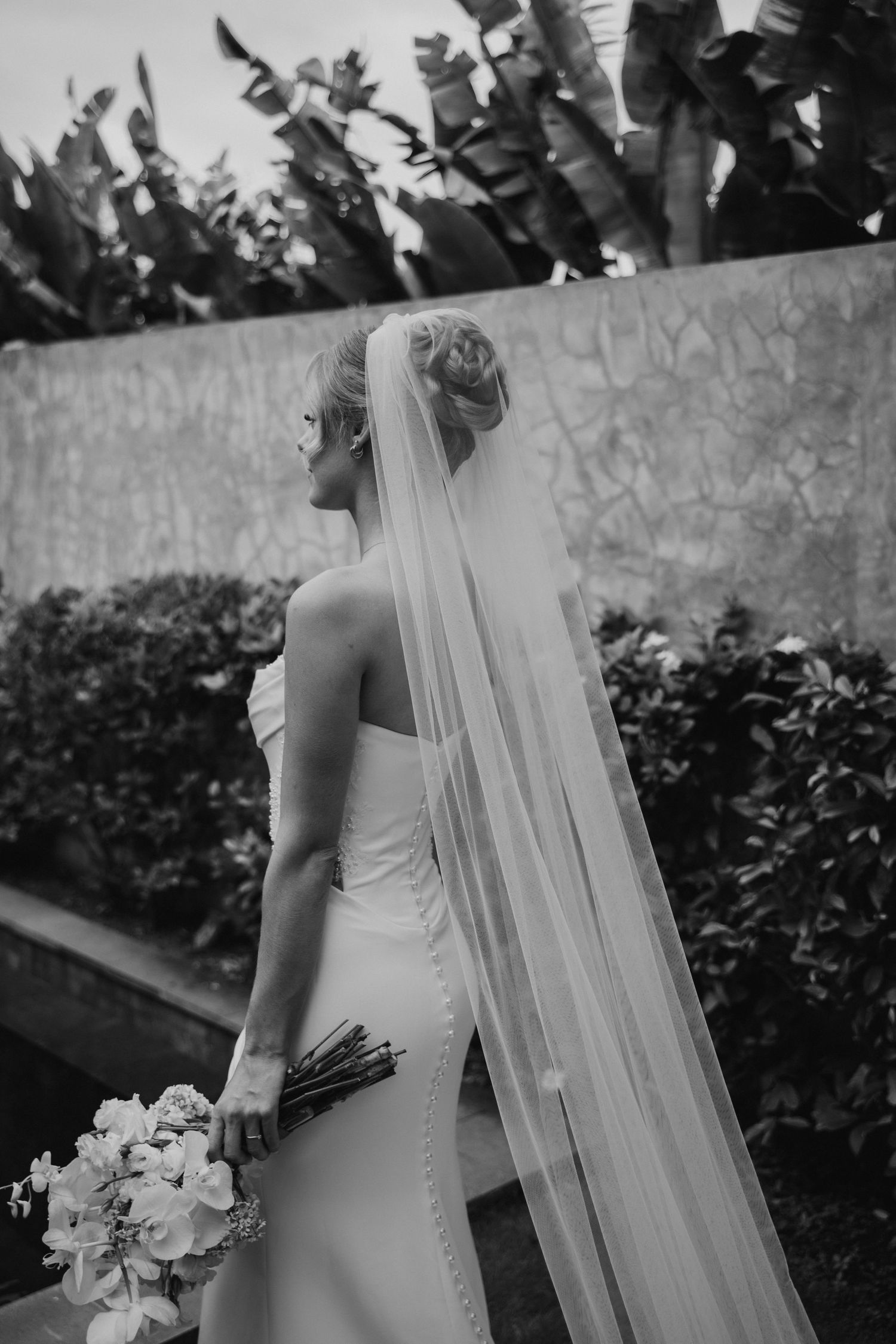 Black and white portrait of bride in fitted gown and long veil holding bouquet outdoors.