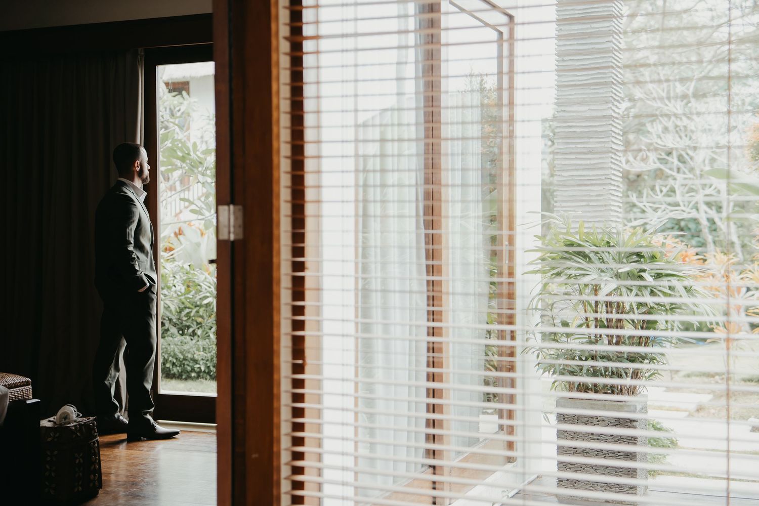 Groom in suit standing by window with venetian blinds on wedding morning.