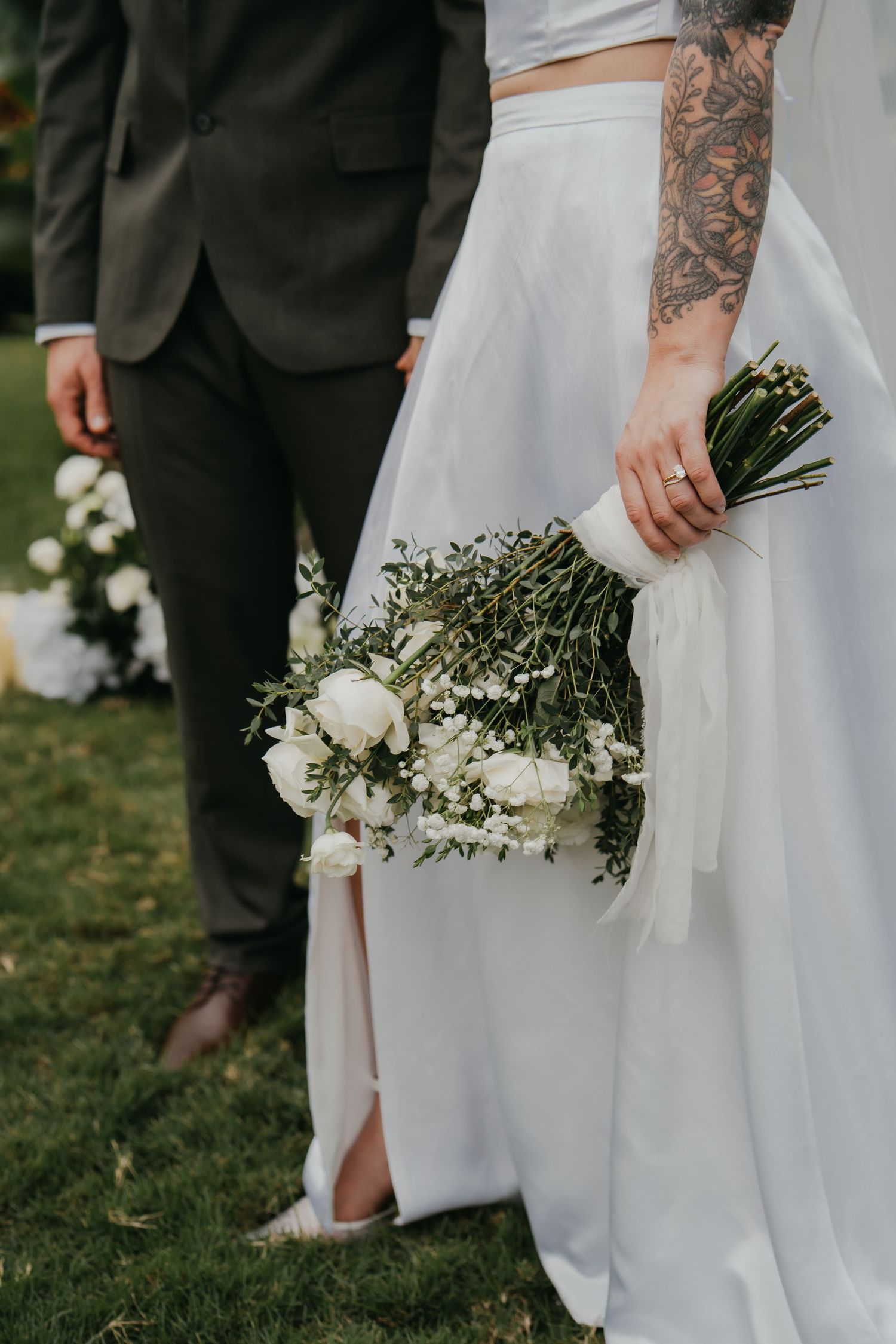 Tattooed bride holding white rose bouquet with ribbon in garden wedding.
