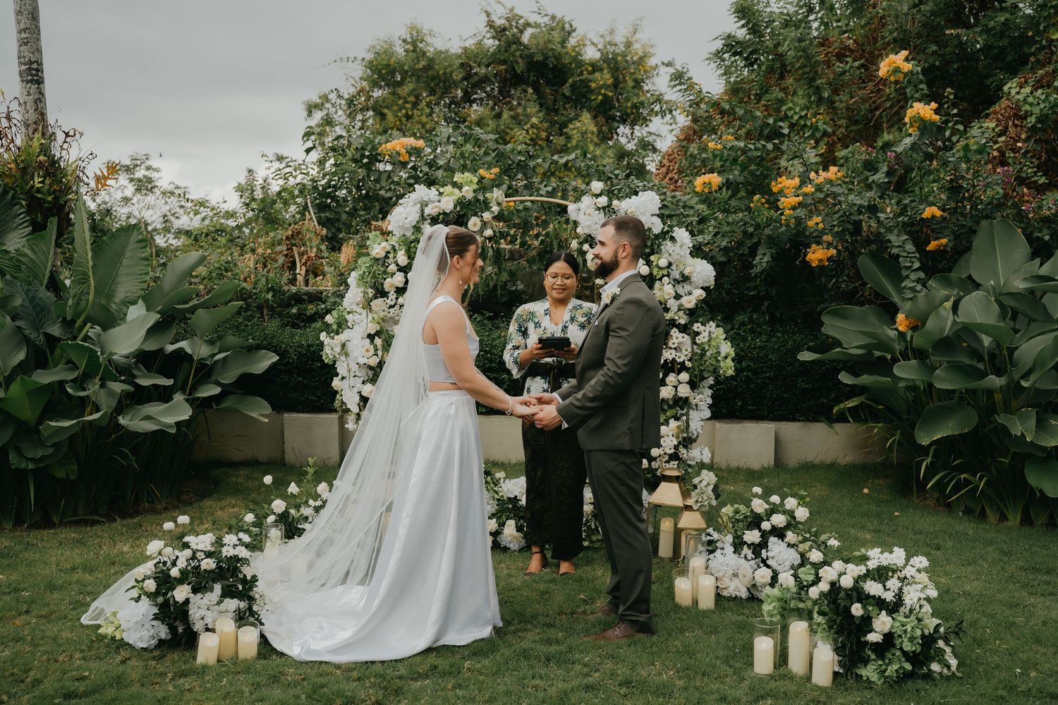 Bride and groom exchanging vows under white floral arch with candles and officiant.